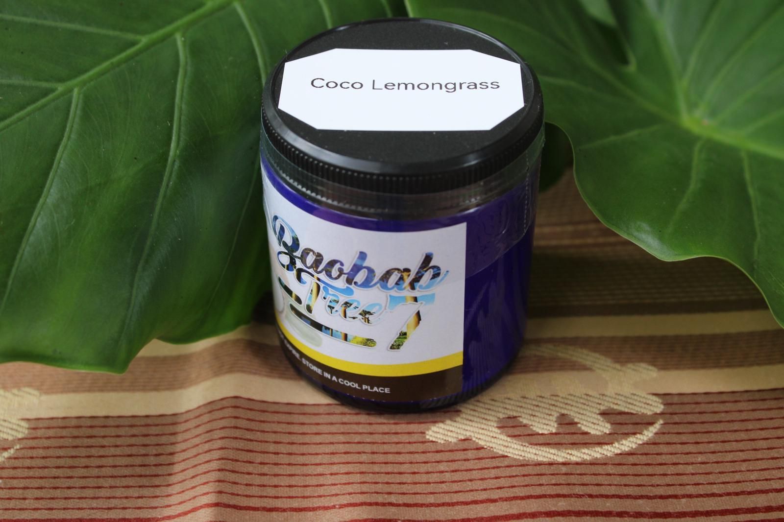 Jar of coconut lemongrass balm on woven mat, framed by green leaves.