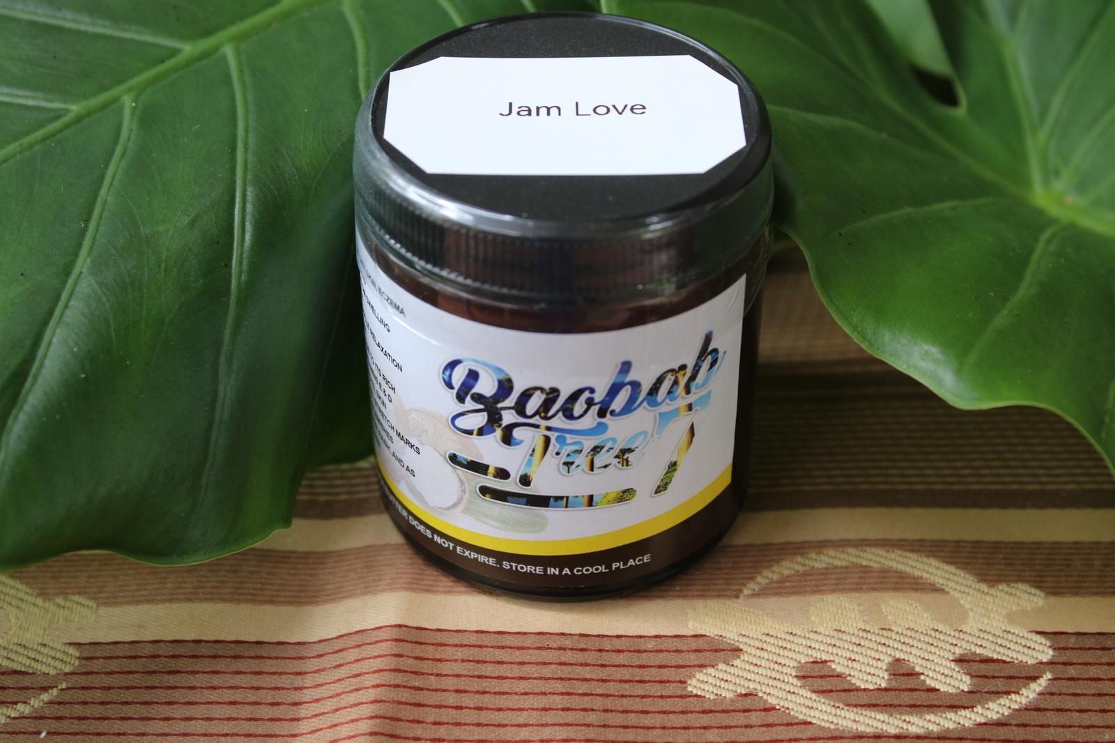 Jar of jam with a white lid on a patterned mat, backed by large green leaves.
