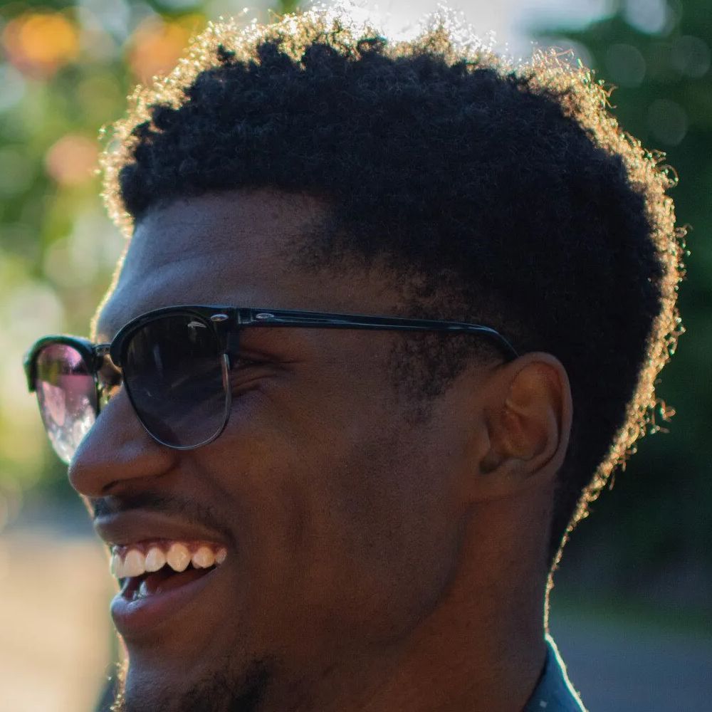 Smiling person in sunglasses outdoors, with sunlit greenery in the background