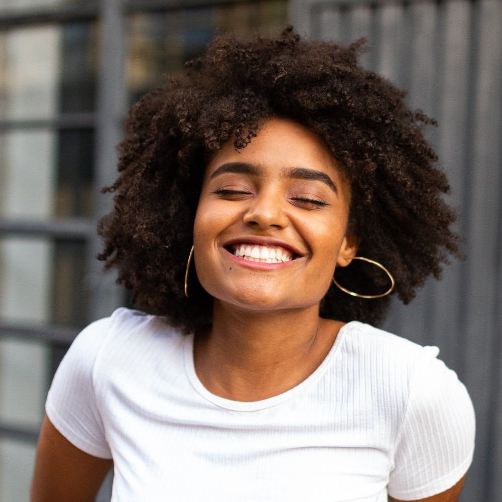Smiling woman with curly hair in a white shirt against a blurred urban background