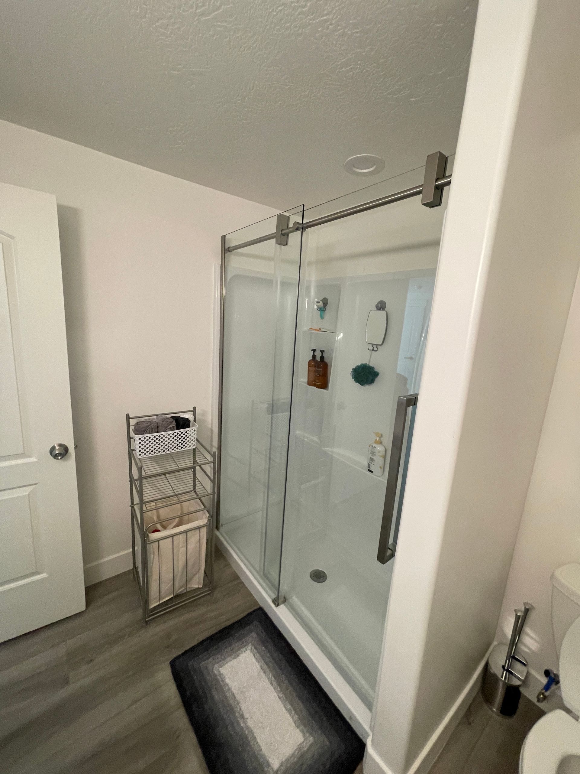 Small bathroom with a glass shower, dark bath mat, and towel rack beside a white door.