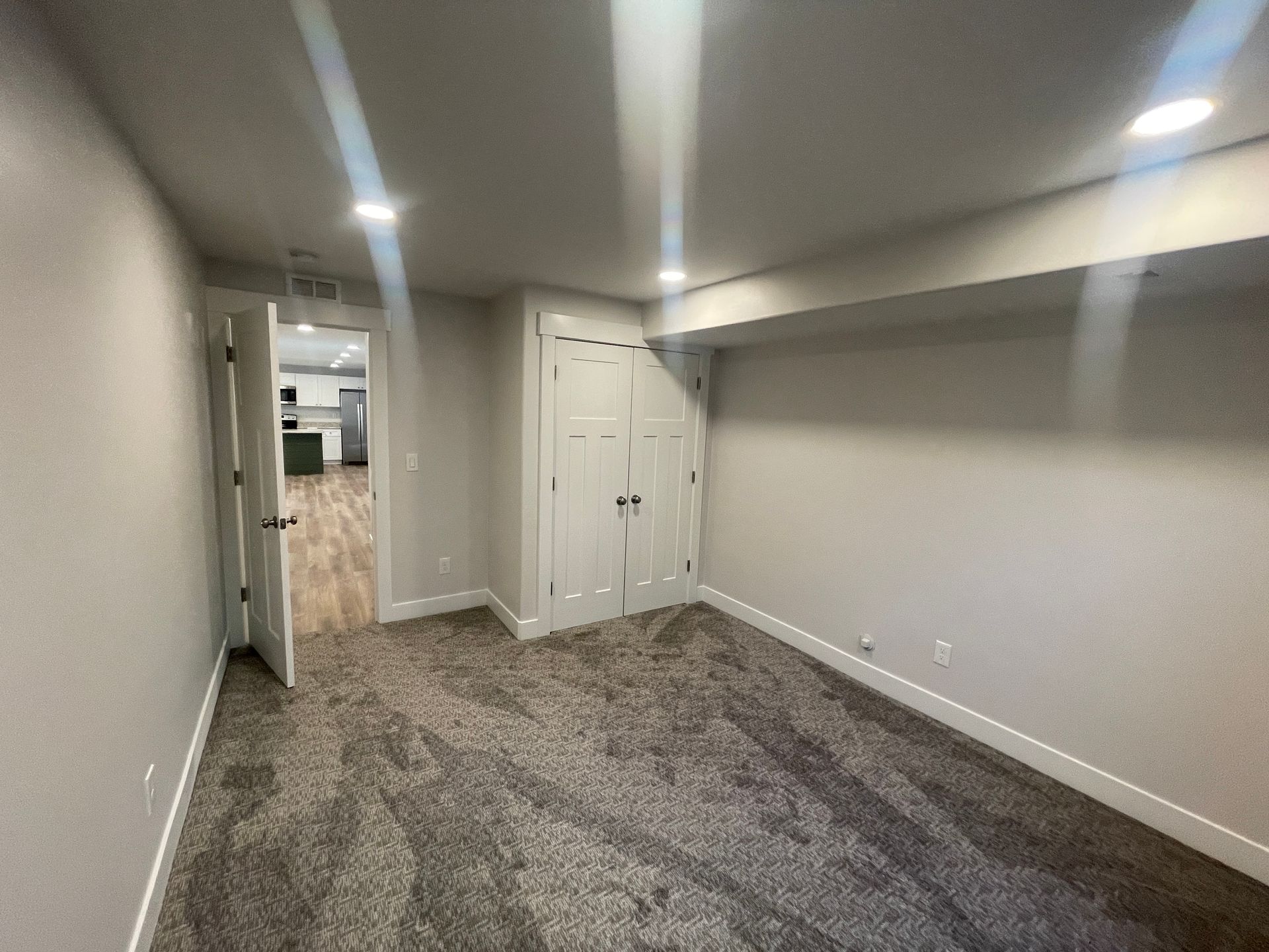 An empty room with a carpeted floor and a door leading to a kitchen.