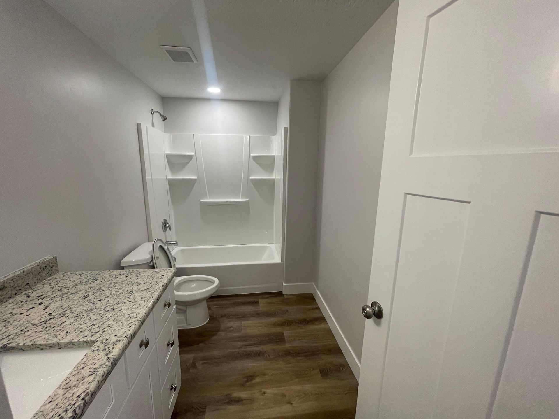 Small bathroom with white built-in shelves, granite vanity, toilet, and wood-look floor.