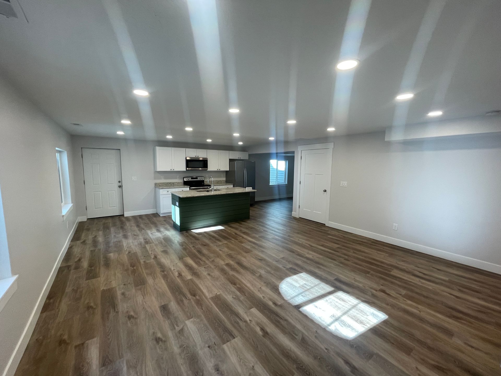 Empty modern living room with wood floors, white walls, recessed lights, and a kitchen island in back
