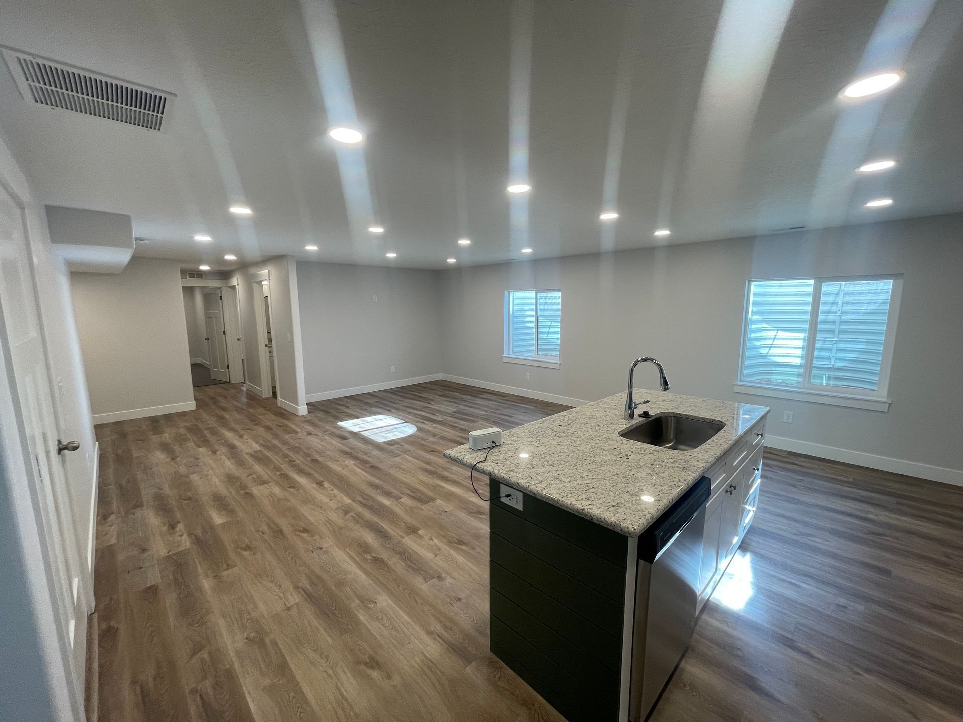 Empty modern kitchen with wood floors, white walls, island sink, and large windows with blinds