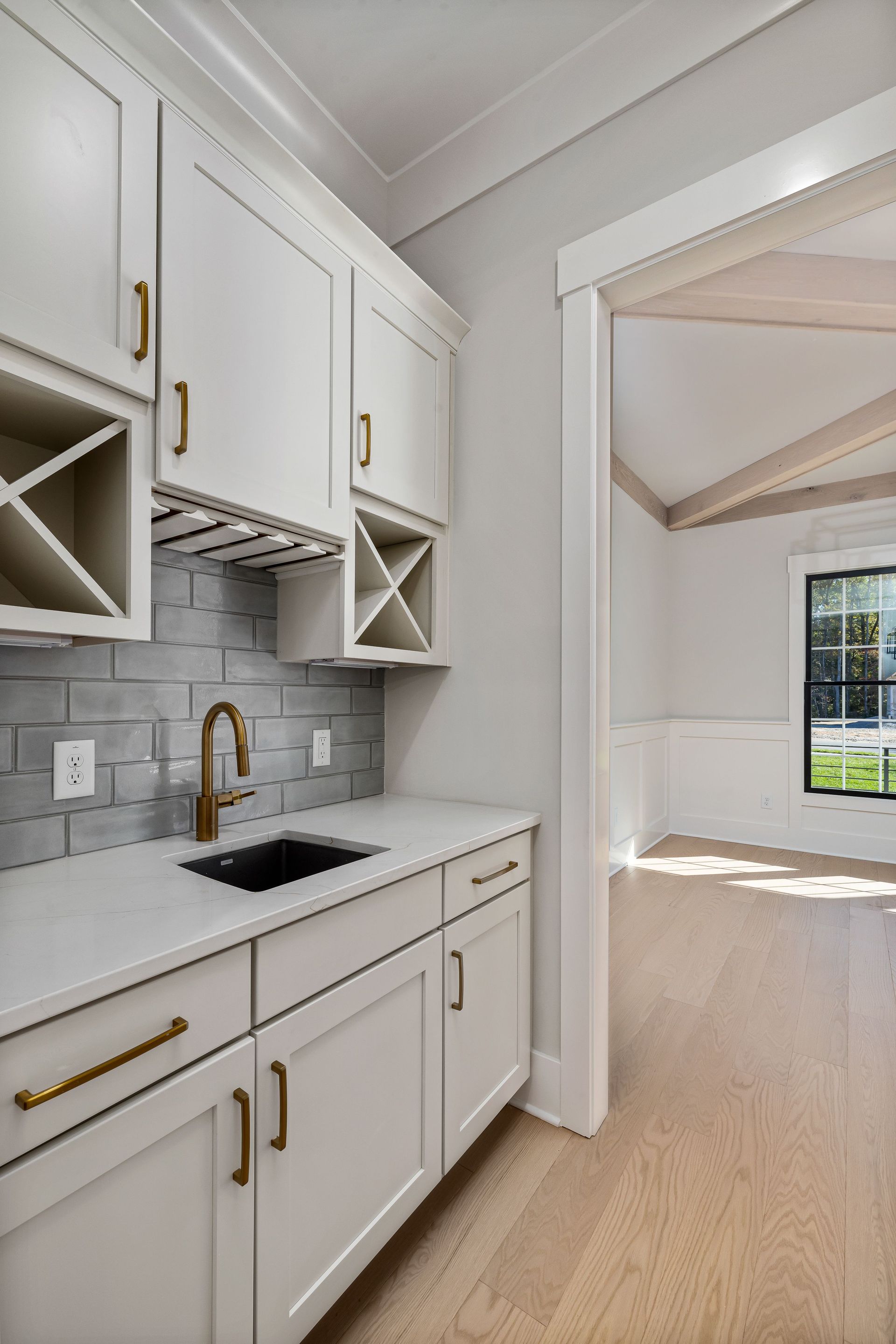 Bright white kitchen with gold accents, black sink, and an open view into a sunlit room with wood floors