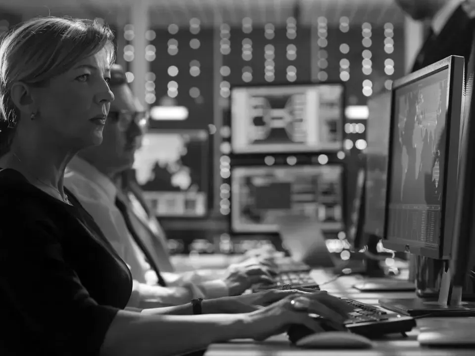 Two people at computer monitors in a dim control room, typing amid multiple screens and data displays