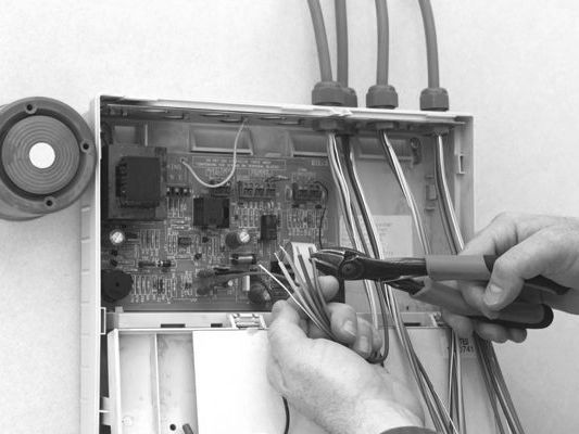 Technician wiring an electrical control box with a screwdriver, exposed circuits and cables visible.
