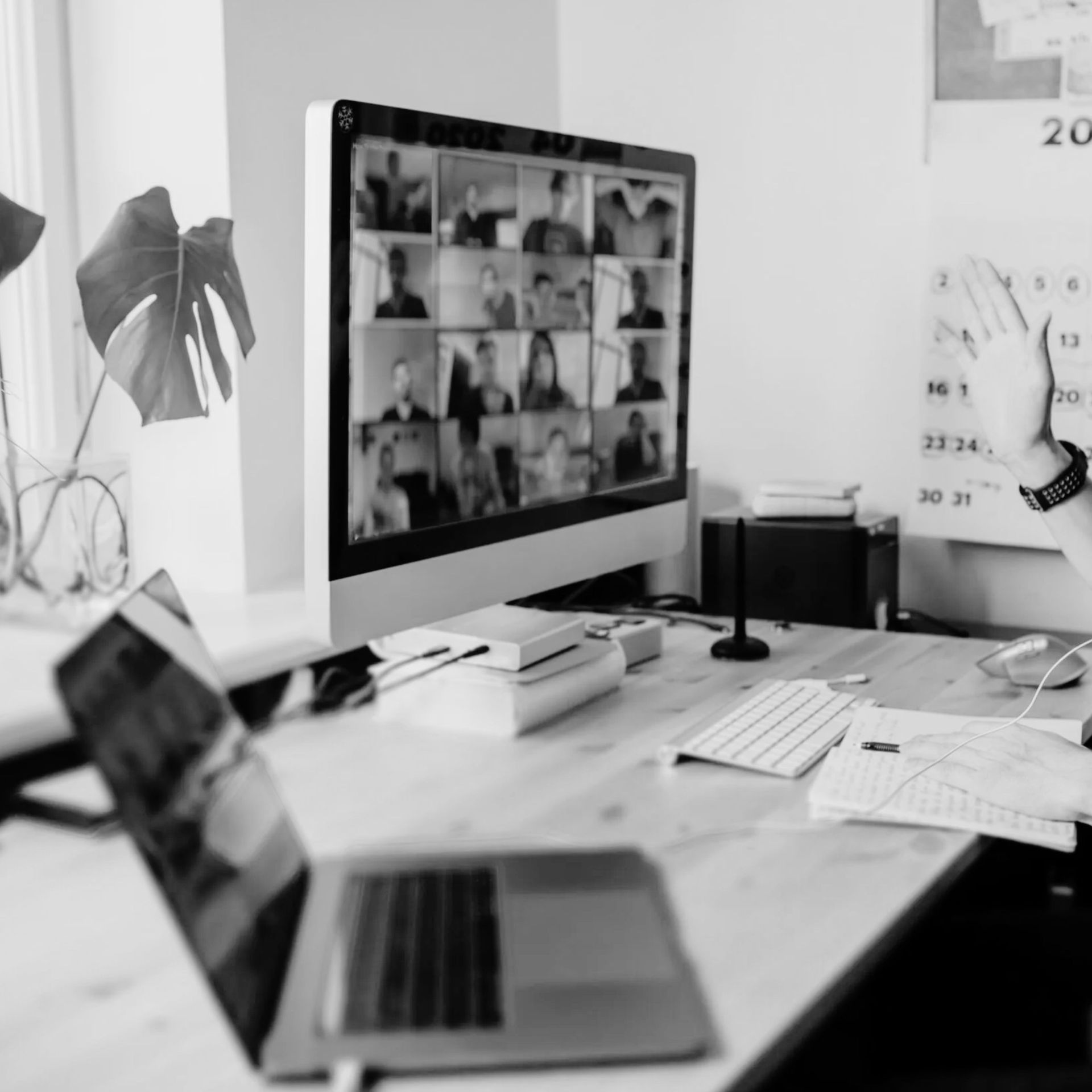 Black-and-white home office with laptop, desktop video call, desk papers, and calendar in the background