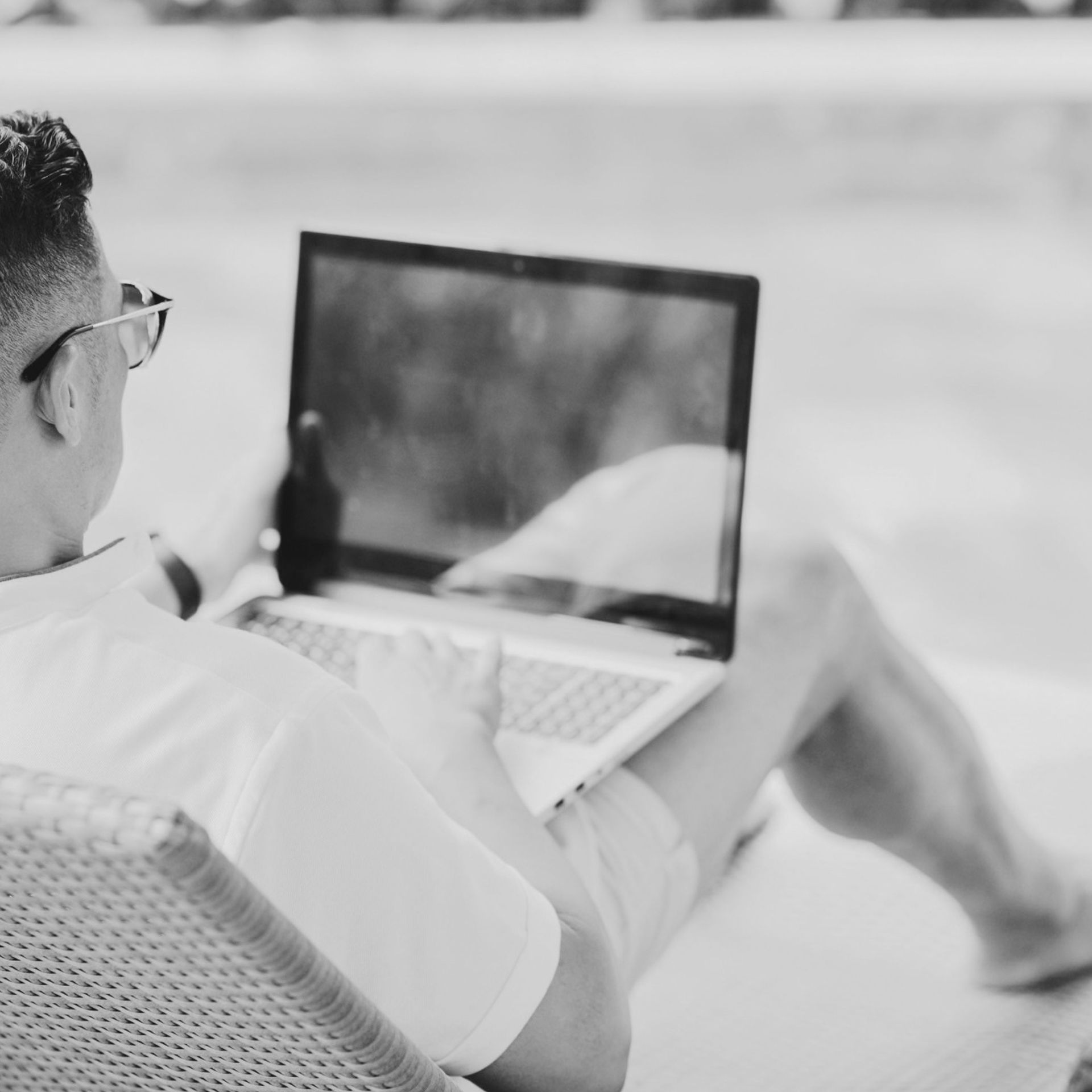 Person sitting outdoors using a laptop on a wicker chair, seen from behind in black and white