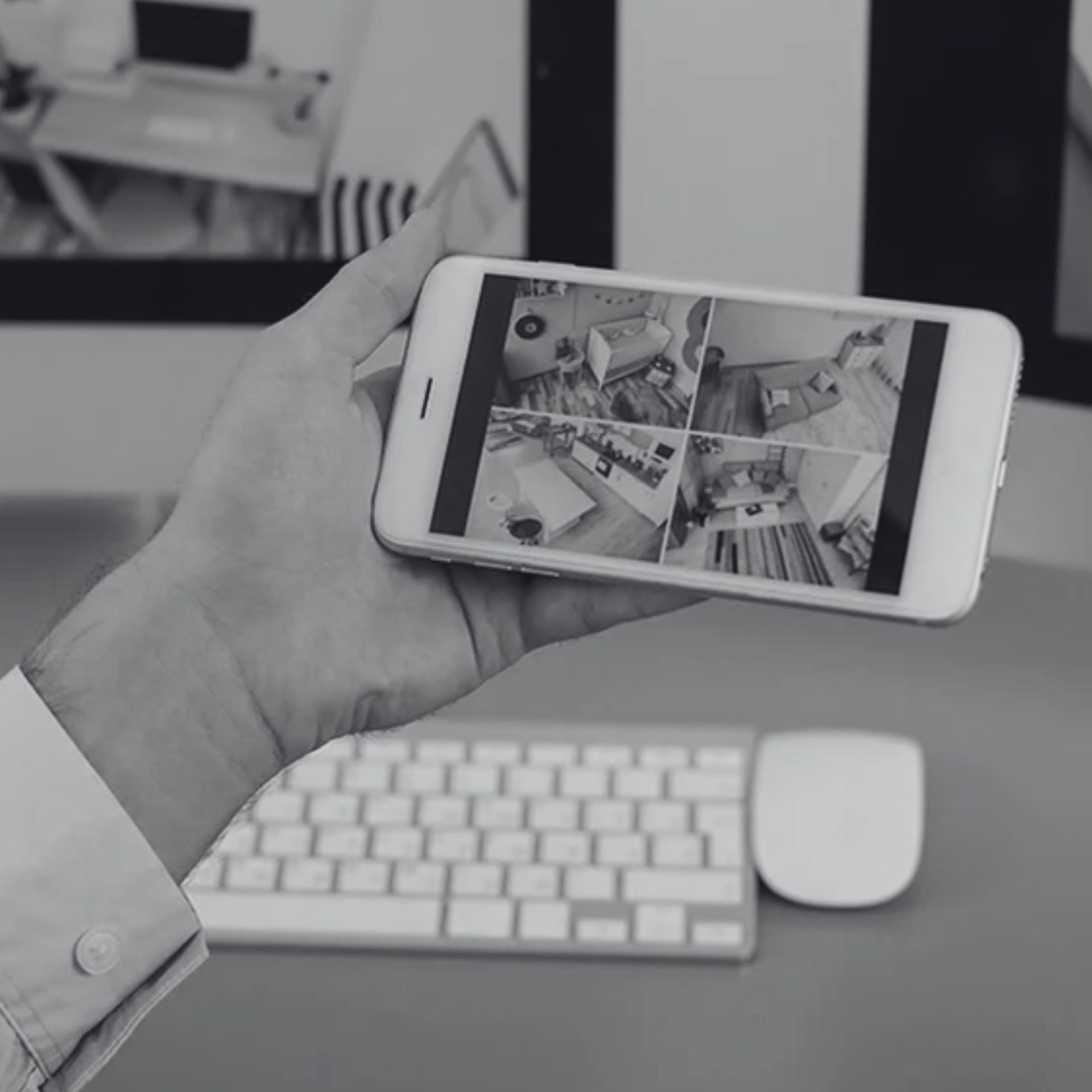 Hand holding a smartphone showing security camera feeds above a desk with keyboard and mouse.