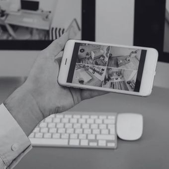 Hand holding a smartphone showing security camera feeds above a desk with keyboard and mouse.