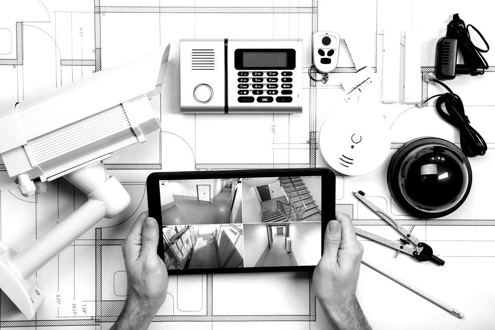 Smart home devices and security items on a blueprint-style desk, with a hand holding a tablet showing a security camera feed