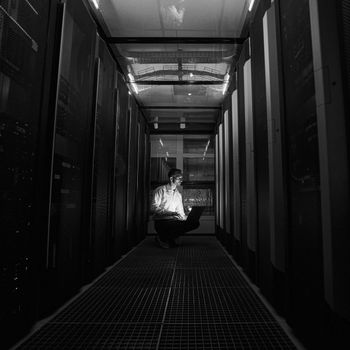 Person seated in a dark server room corridor, illuminated by a bright monitor glow inspecting security systems