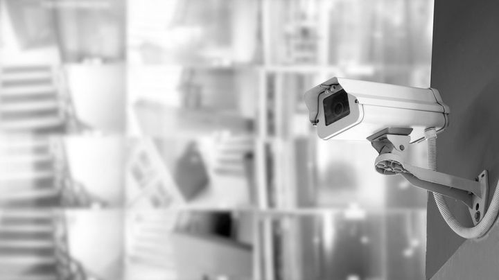 White security camera mounted on a wall, facing left in a blurred indoor office setting