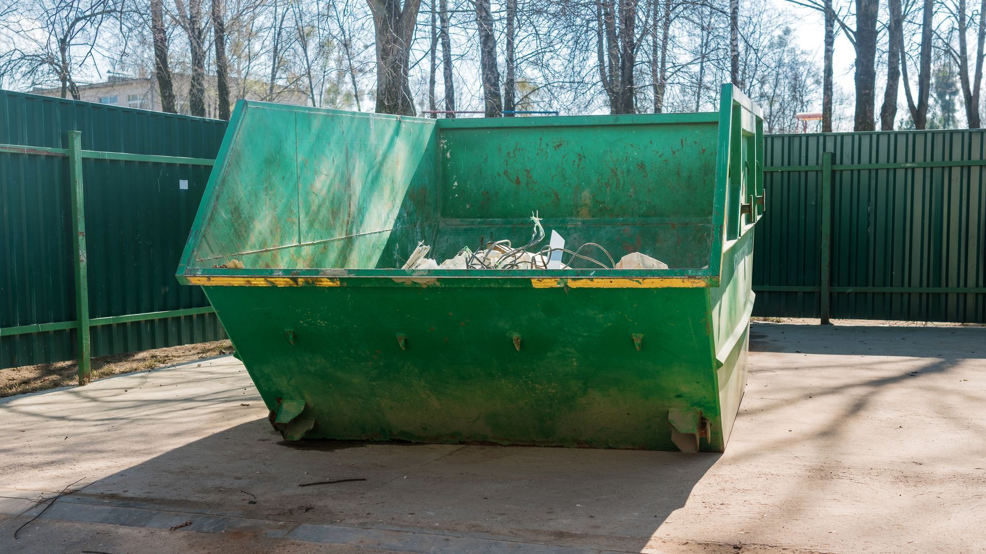 Green dumpster filled with trash sits on a concrete pad with a fence and trees in the background. Green dumpster filled with trash sits on a concrete pad with a fence and trees in the background.