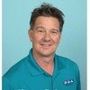 Man with short, dark hair, wearing a teal polo shirt, smiles in front of a blue background.