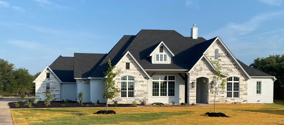 Large stone suburban house with a dark roof and white trim on a sunny lawn