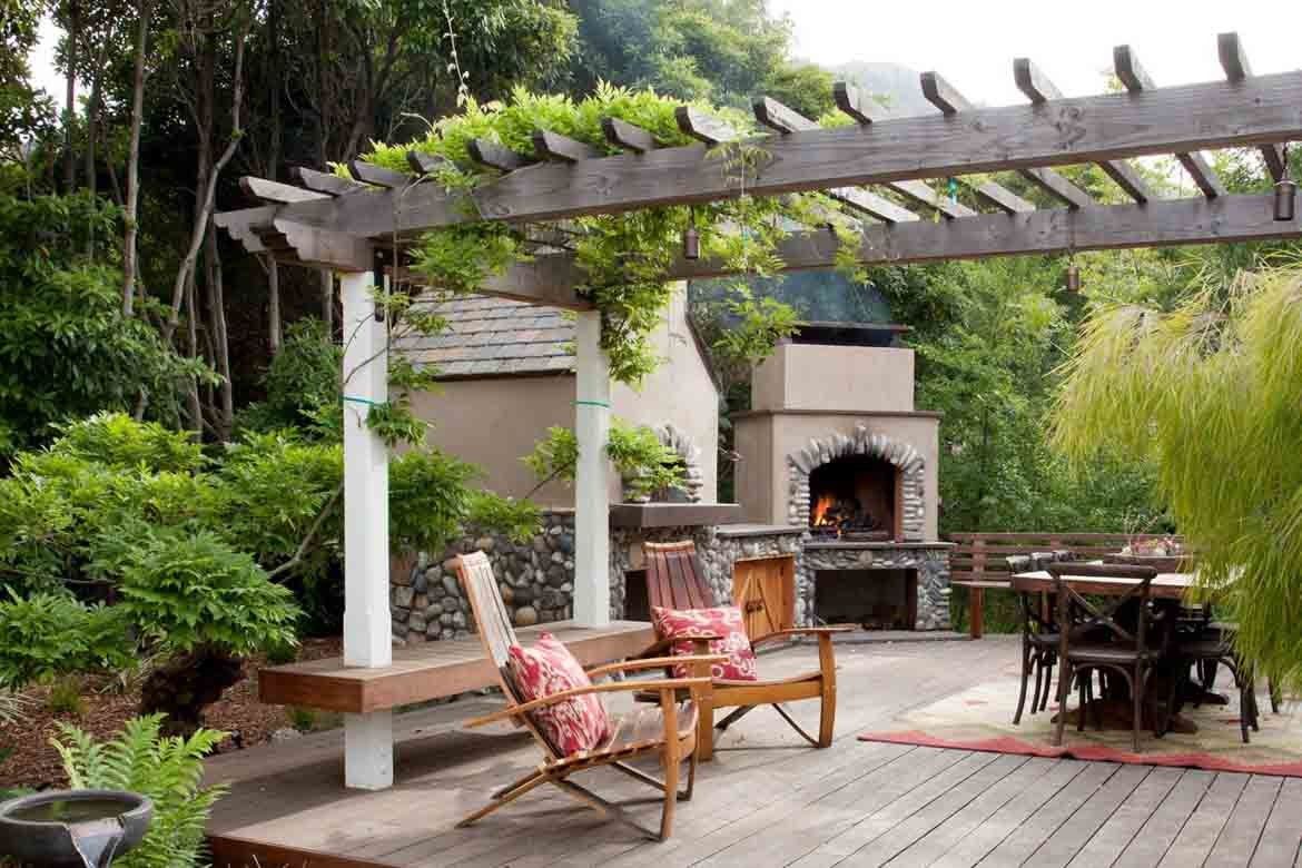 Outdoor patio with wooden pergola, fireplace, lounge chair, and dining table surrounded by greenery