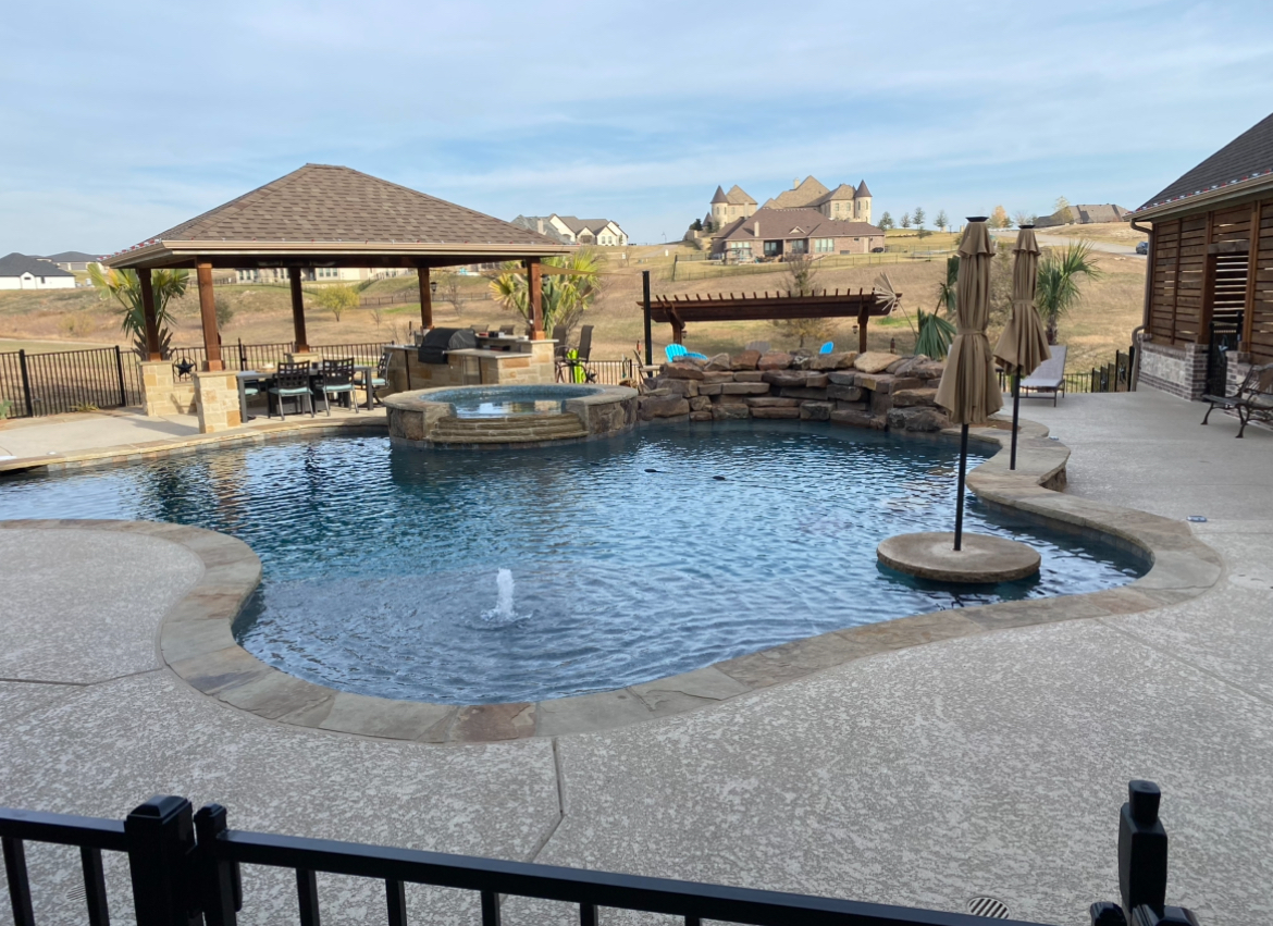 A backyard swimming pool with a stone waterfall, a covered patio, and a water feature under a clear blue sky.