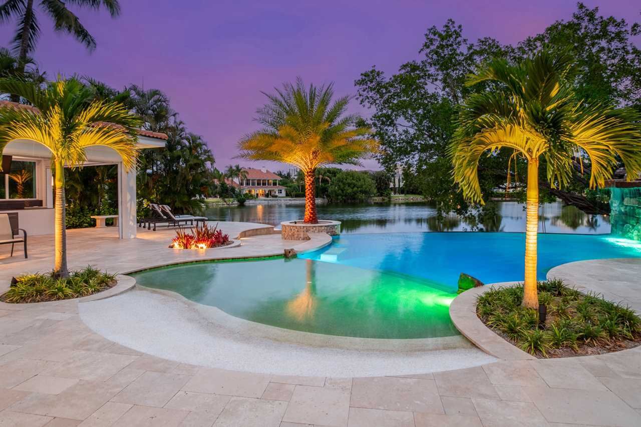 A tranquil pool terrace at sunset, featuring palm trees, a blue infinity-style pool, and a waterfront home in the distance.