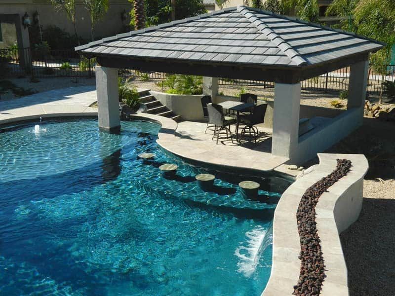 A swimming pool with an underwater bar, built-in stools, and a covered patio area with a table and chairs.