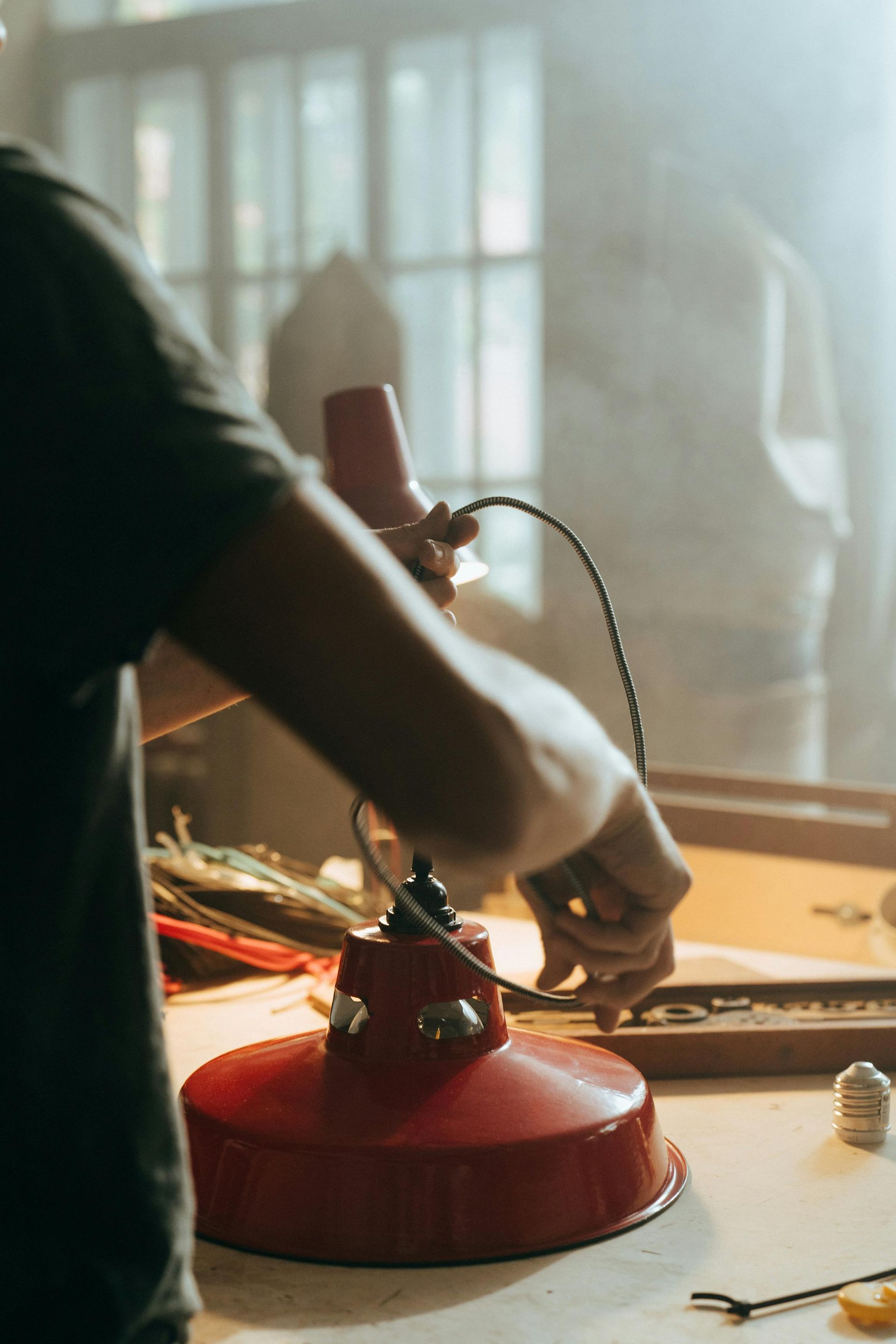 Person repairing a red lamp on a workbench in a sunlit workshop