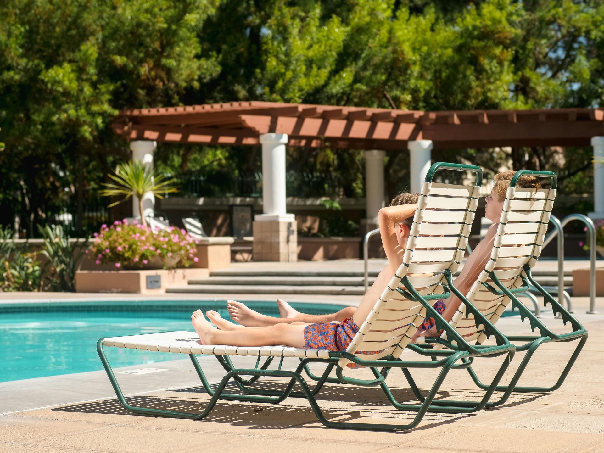 Two lounge chairs by a pool under trees and a pergola, with a small flower bed nearby