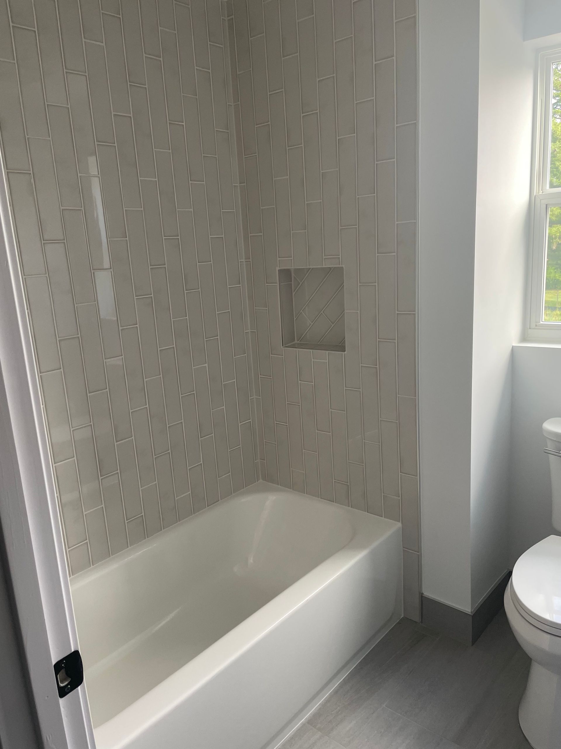 Bathroom with white bathtub, tiled shower wall, small niche, and partial toilet visible near a window.