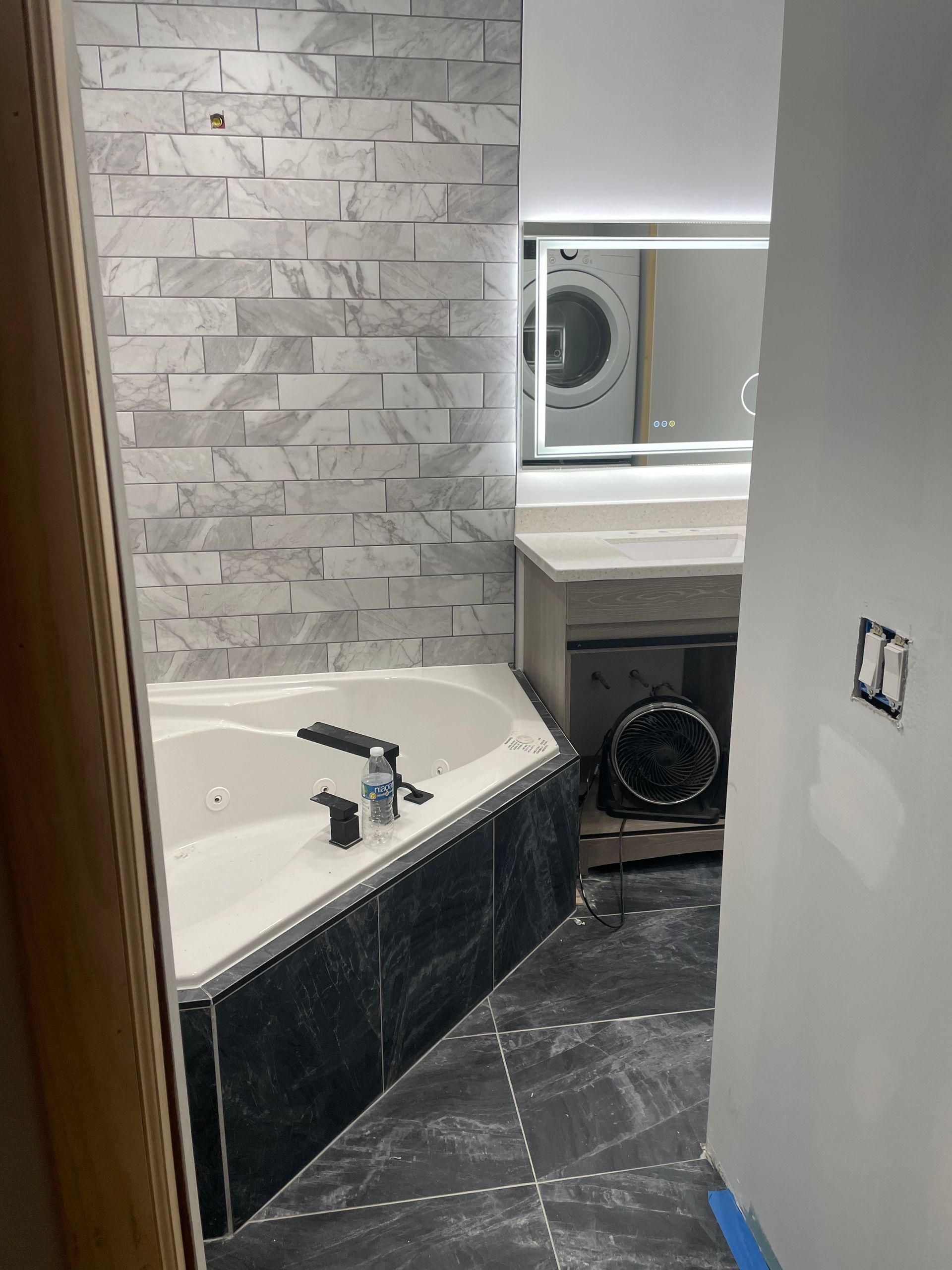 Bathroom with a large tub, gray tile walls, and a laundry machine visible through an opening