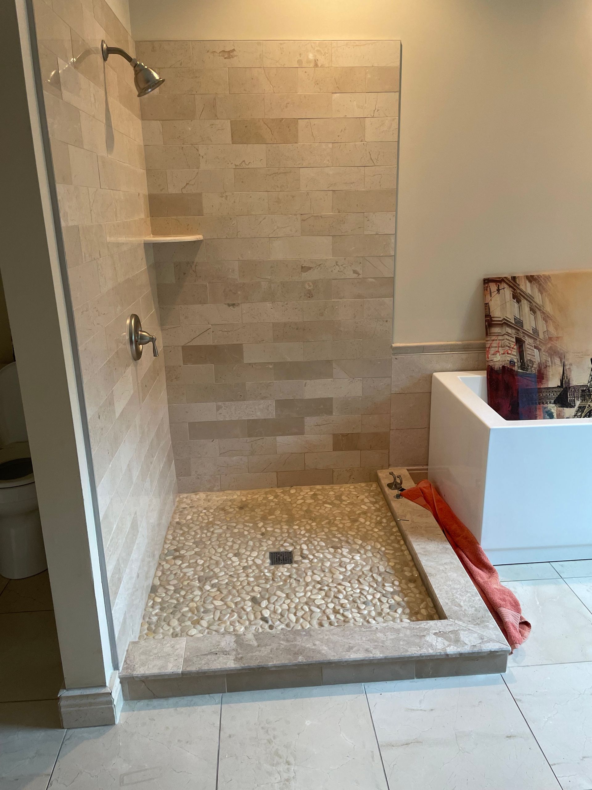Bathroom shower with beige tile, pebble floor, and white tub nearby, under renovation