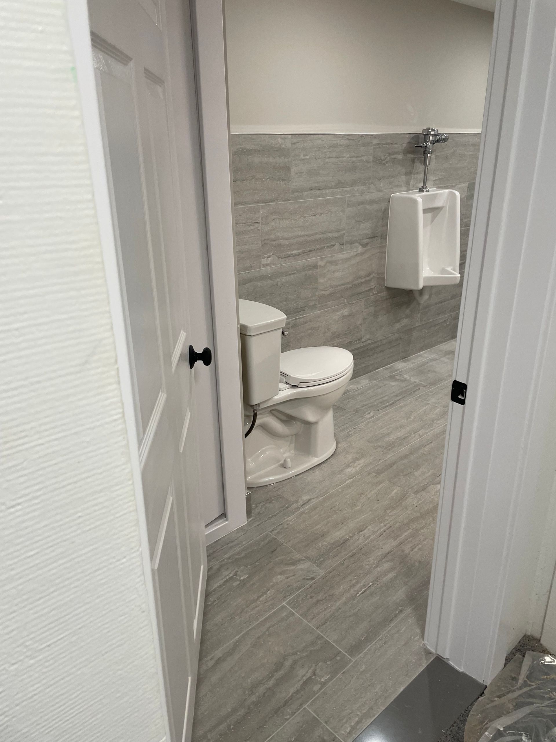 Small bathroom with toilet, wall-mounted paper towel dispenser, and gray tile walls viewed through doorway