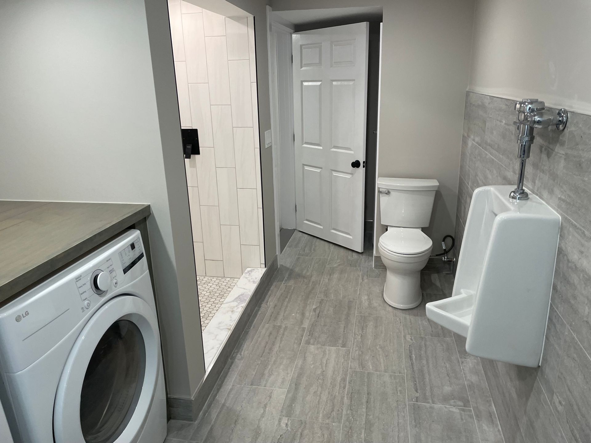Small bathroom with a toilet, urinal, sink, and open door to a shower area, gray flooring and white walls.