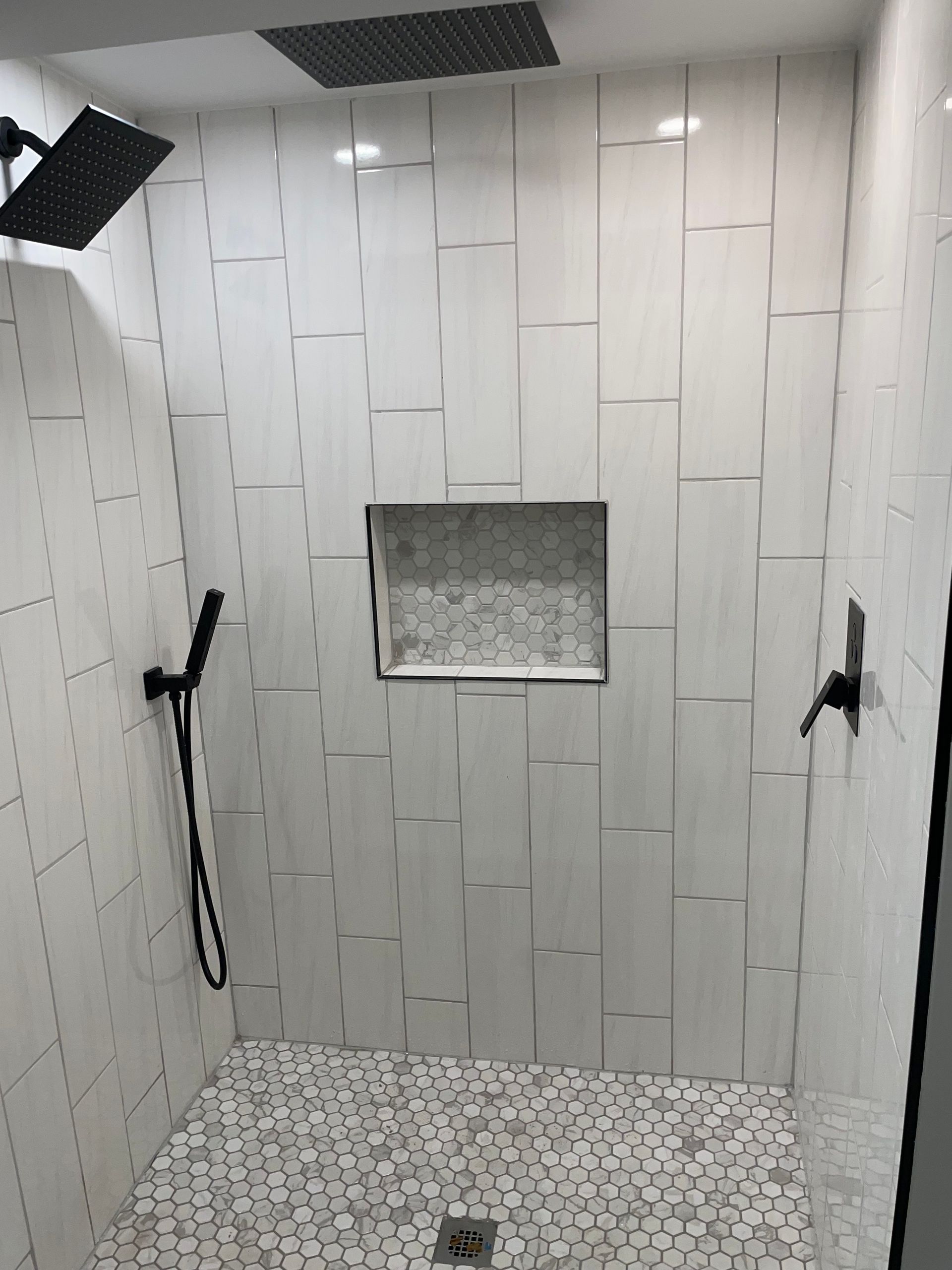 Walk-in shower with gray tiled walls, two black grab bars, a small window, and a mosaic floor.