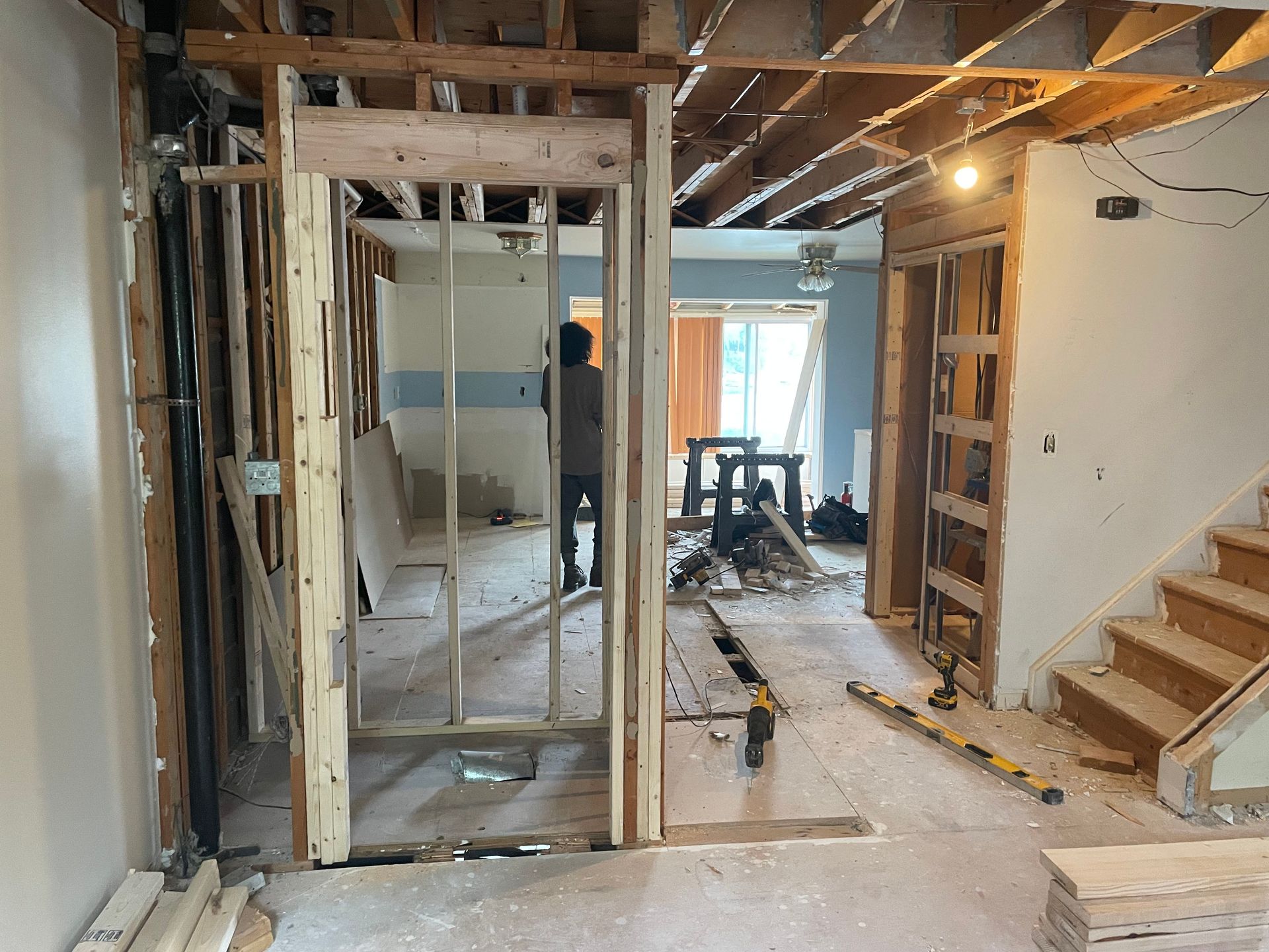 Interior under renovation with exposed framing, tools, stairs, and a hallway opening to a bright room.
