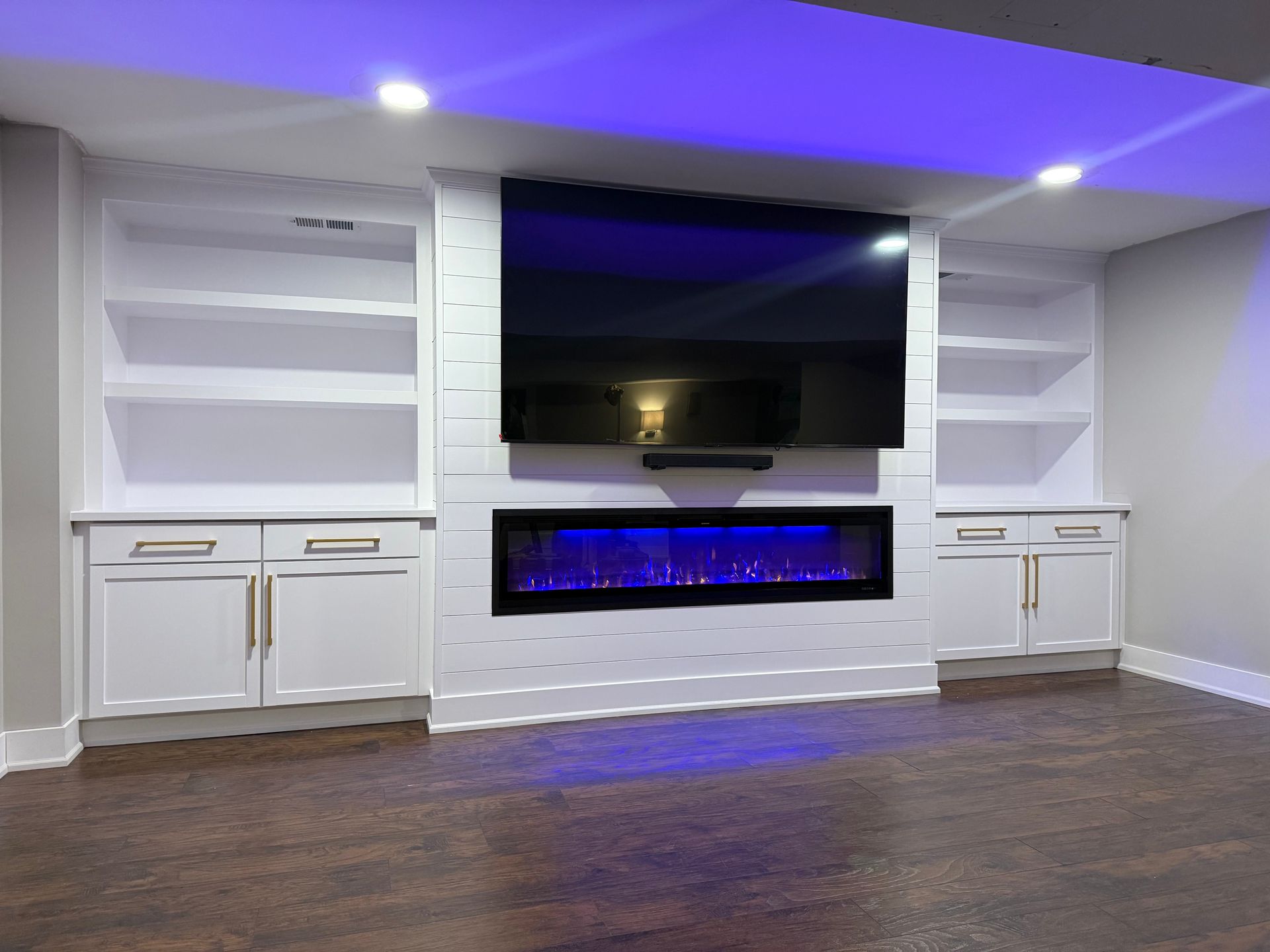 Modern living room wall unit with built-in TV, electric fireplace, and white cabinets under blue accent lighting