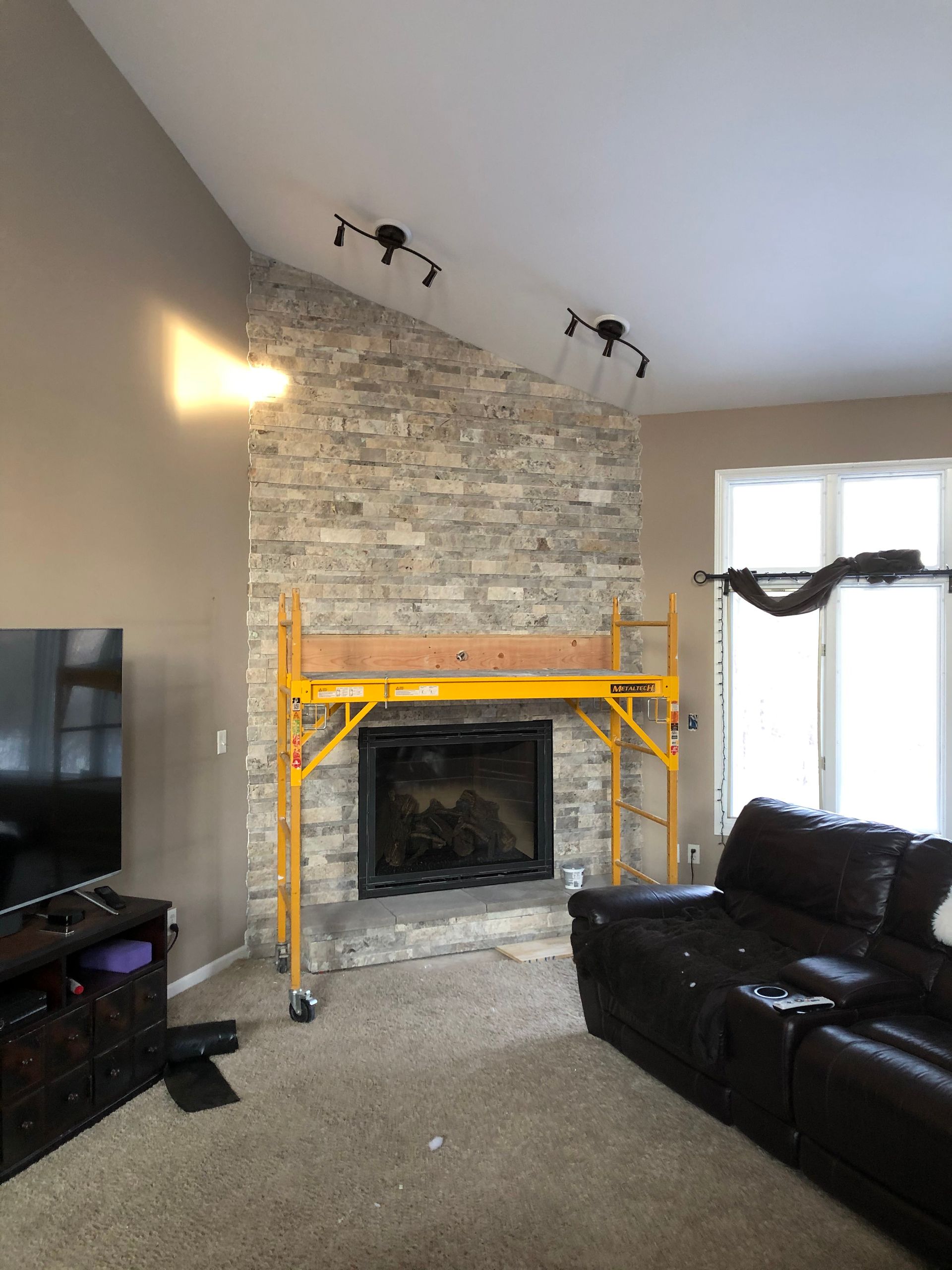 Living room with stone fireplace, yellow scaffold, black leather sofa, TV, and window with black drapes