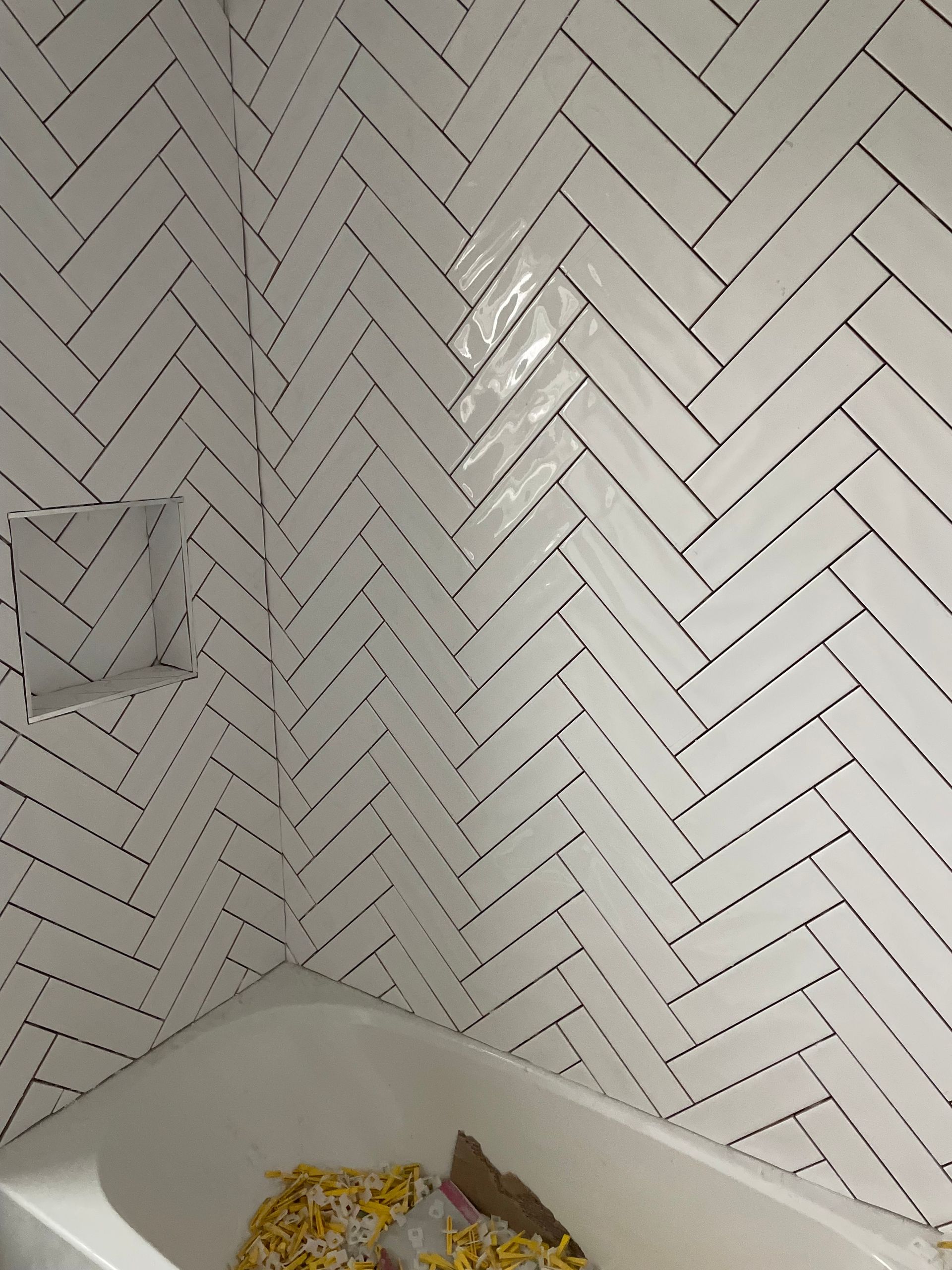 White herringbone tile shower wall above a corner tub with a few leaves on the floor