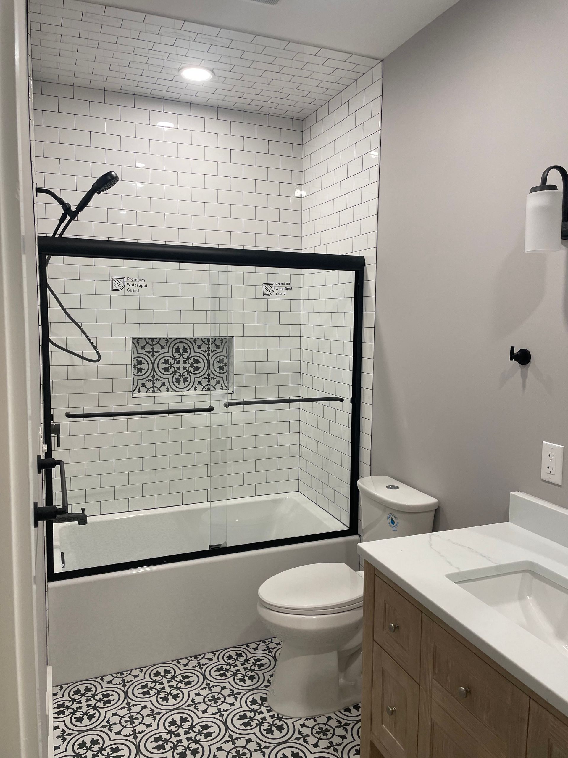 Small bathroom with black-and-white patterned shower, toilet, vanity, and wall sconce.