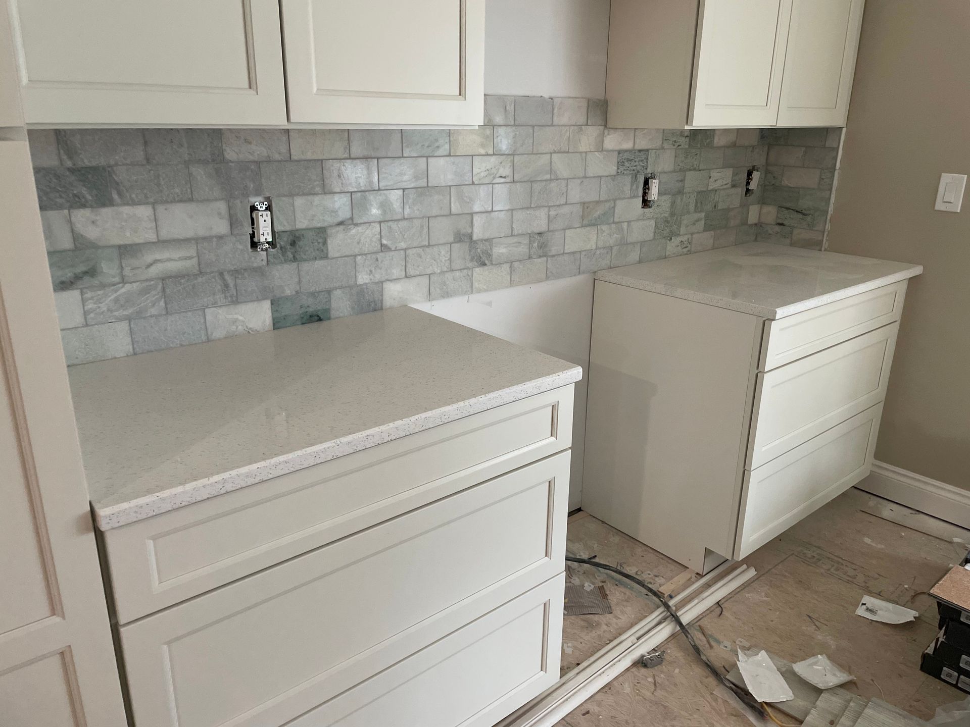 Two white kitchen cabinets installed against a gray tile backsplash in an unfinished kitchen.