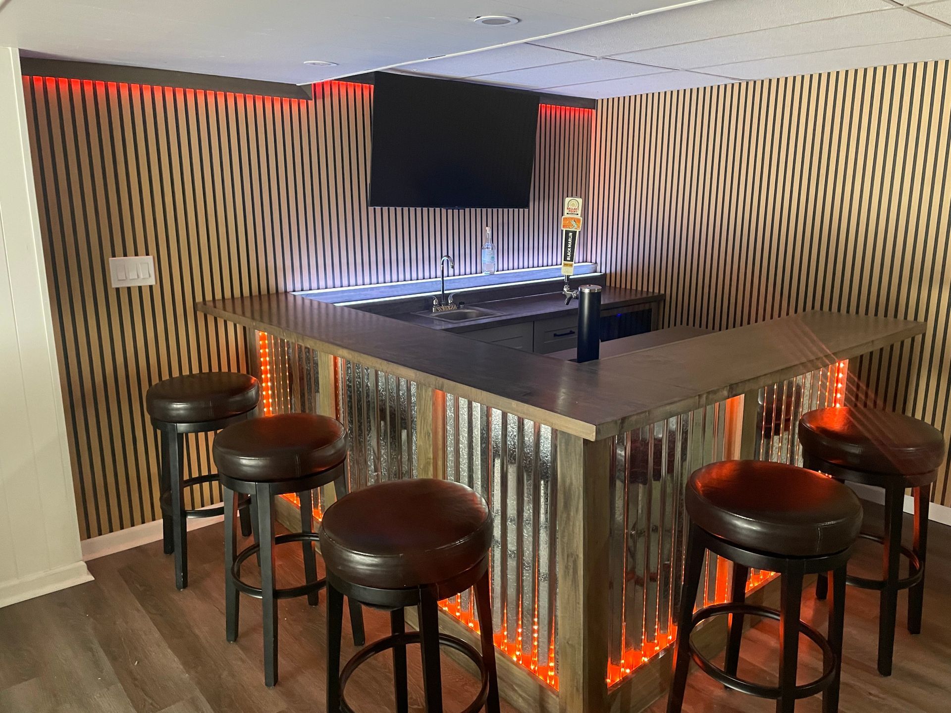 Modern home bar with corrugated metal island, stools, warm orange lighting, and a TV overhead