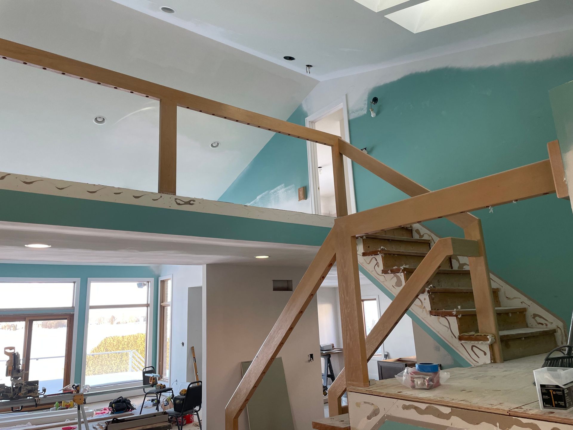 Interior under renovation with wooden stairs, loft railing, and teal walls overlooking a bright room