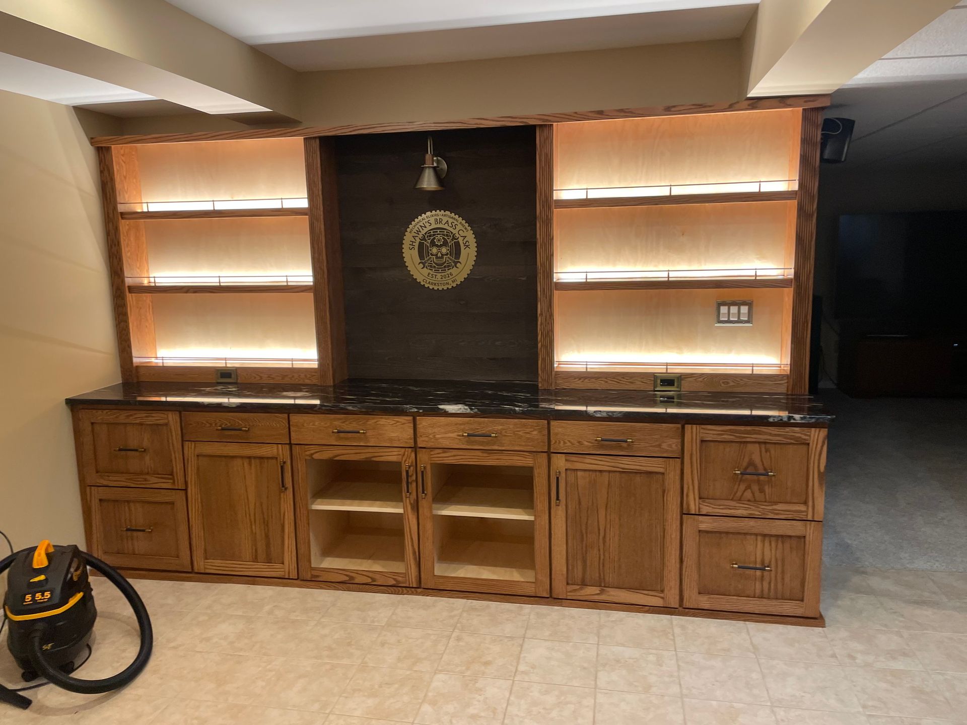 Wooden bar with backlit shelves and a dark stone center panel in a room with a vacuum on the left.