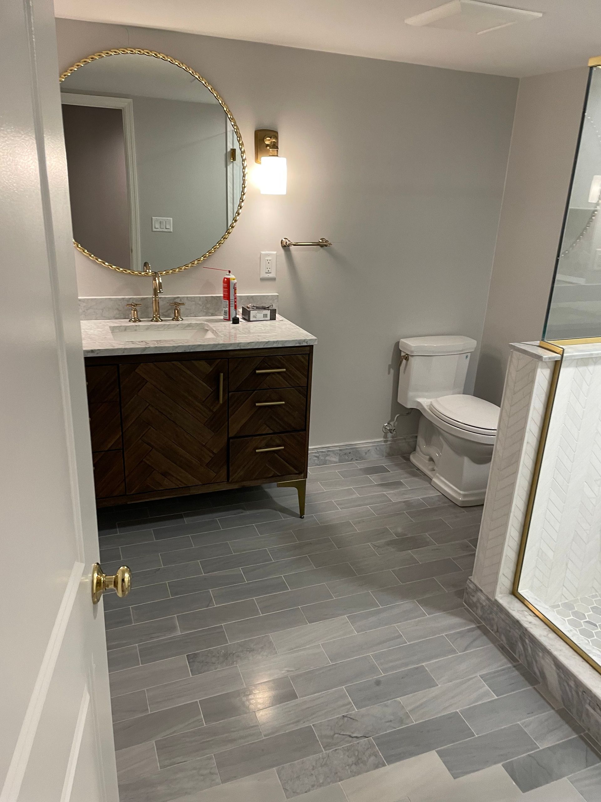 Modern bathroom with gray tile, dark vanity, round mirror, wall sconce, toilet, and glass shower door