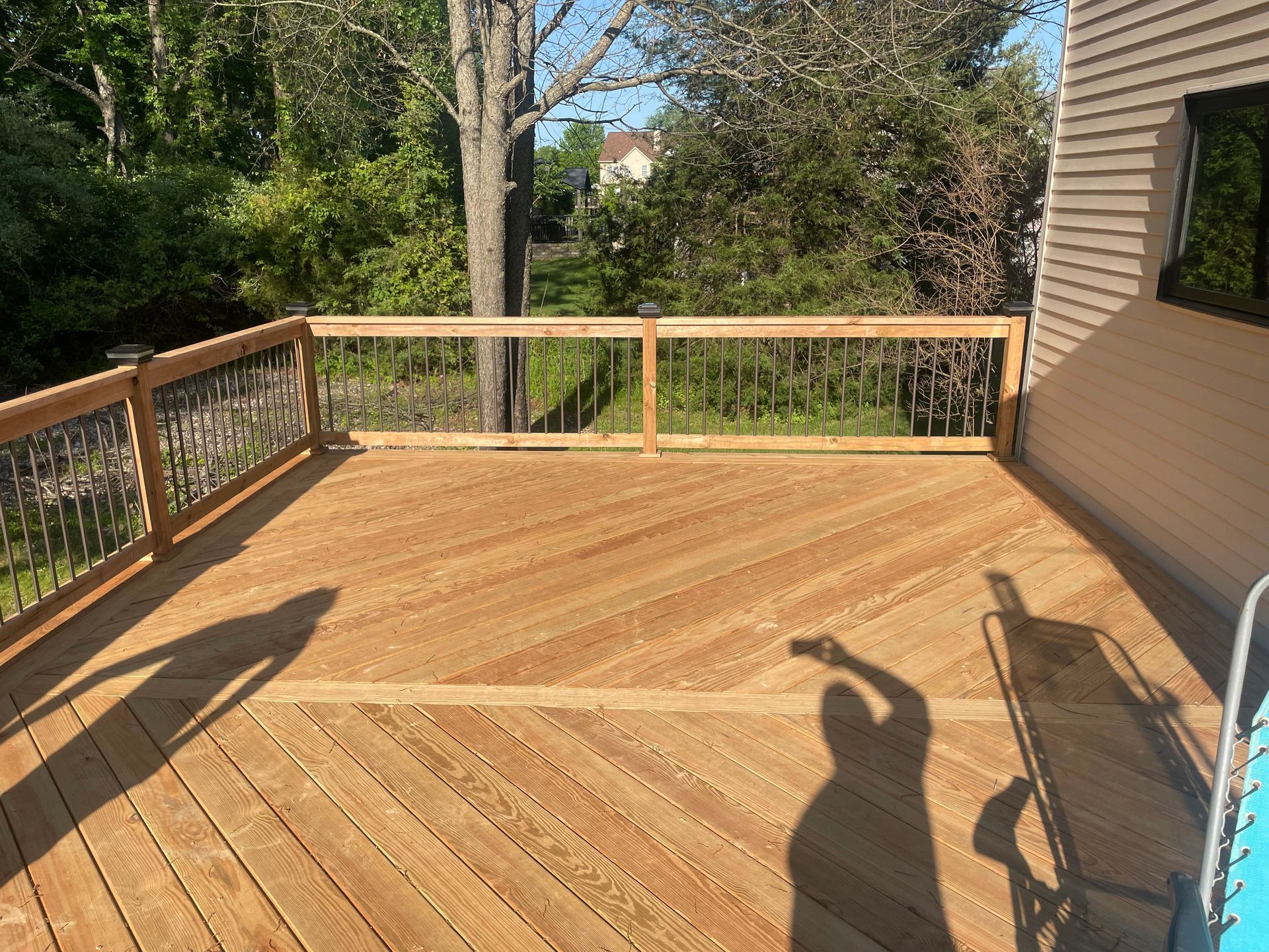 Wooden backyard deck with railings and stairs, sunlight casting long shadows, trees in the background