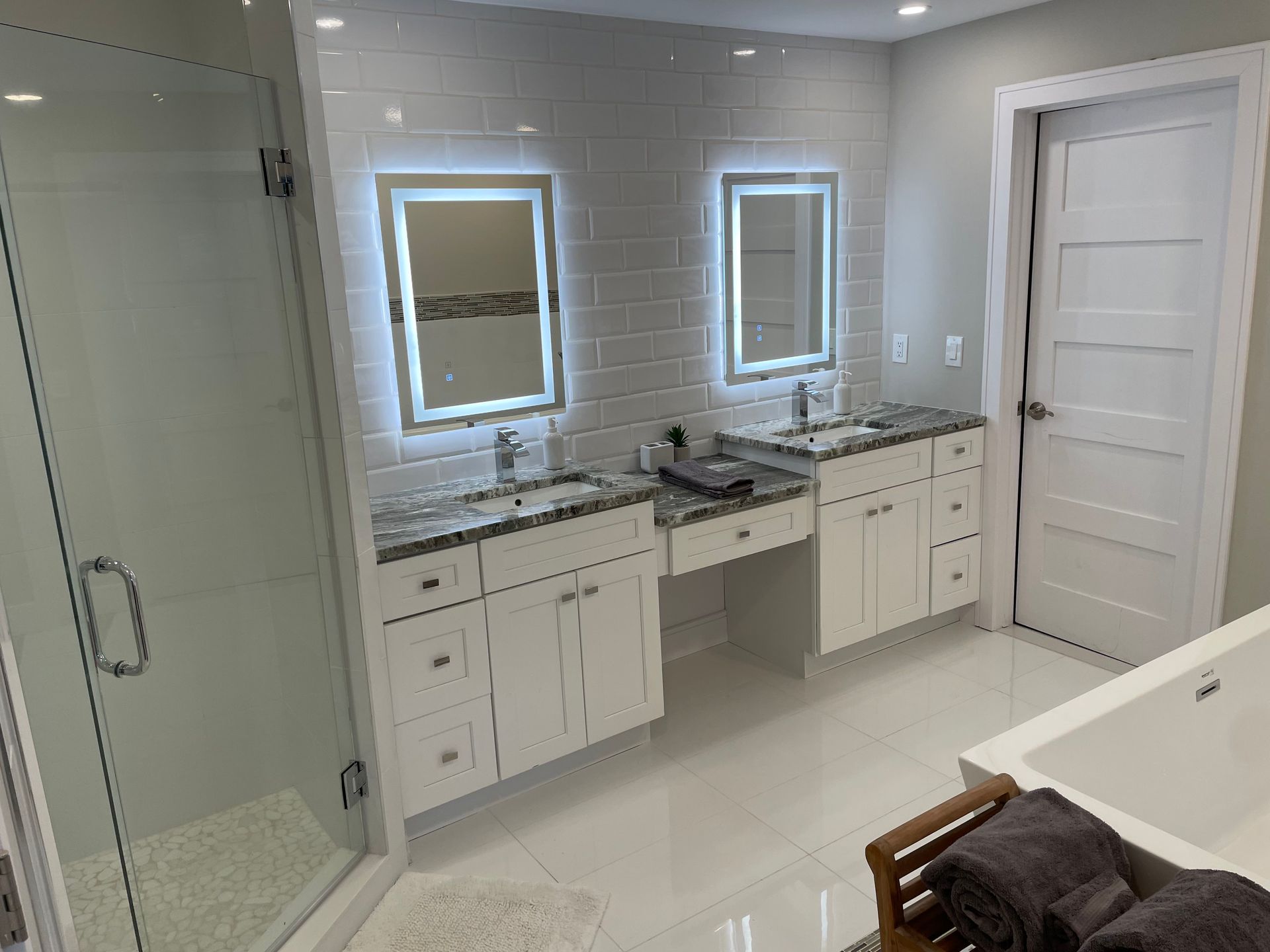 Modern bathroom with glass shower, double vanity, lit mirrors, and white tile walls