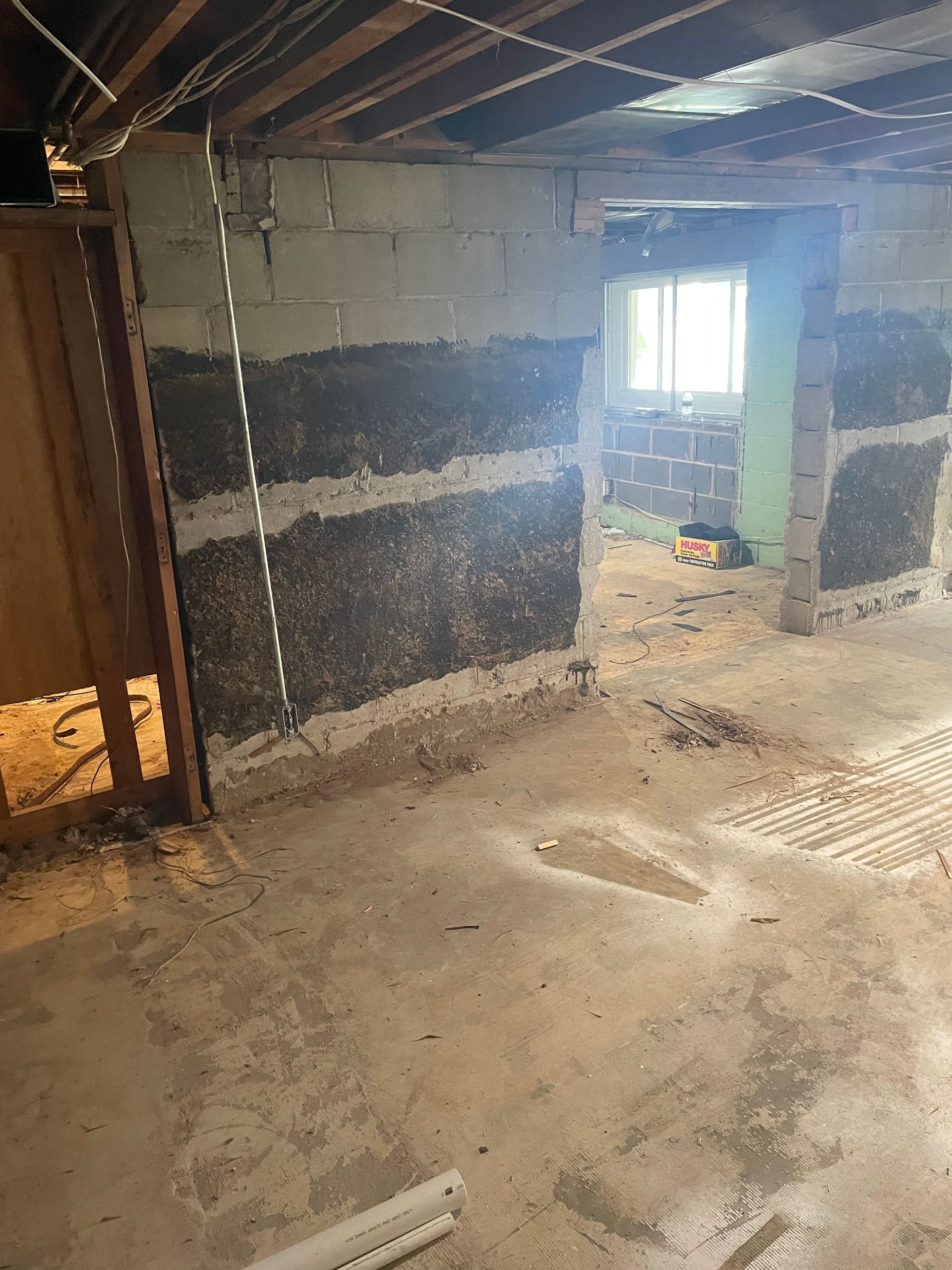 Damaged basement wall with exposed blocks and debris on a concrete floor, lit by a bright window opening.