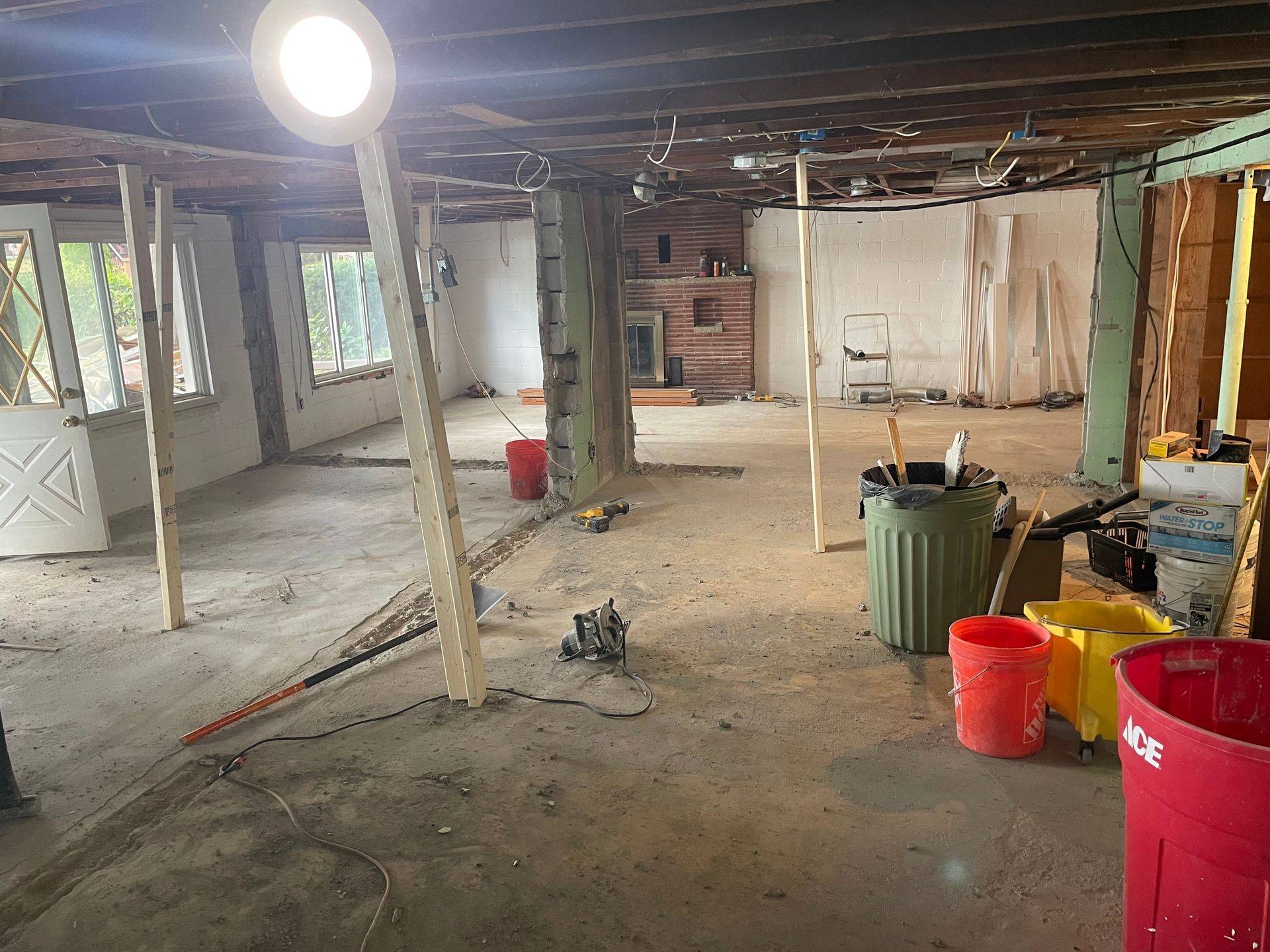 Renovation site with exposed beams, concrete floor, buckets, trash bins, and a bright work light