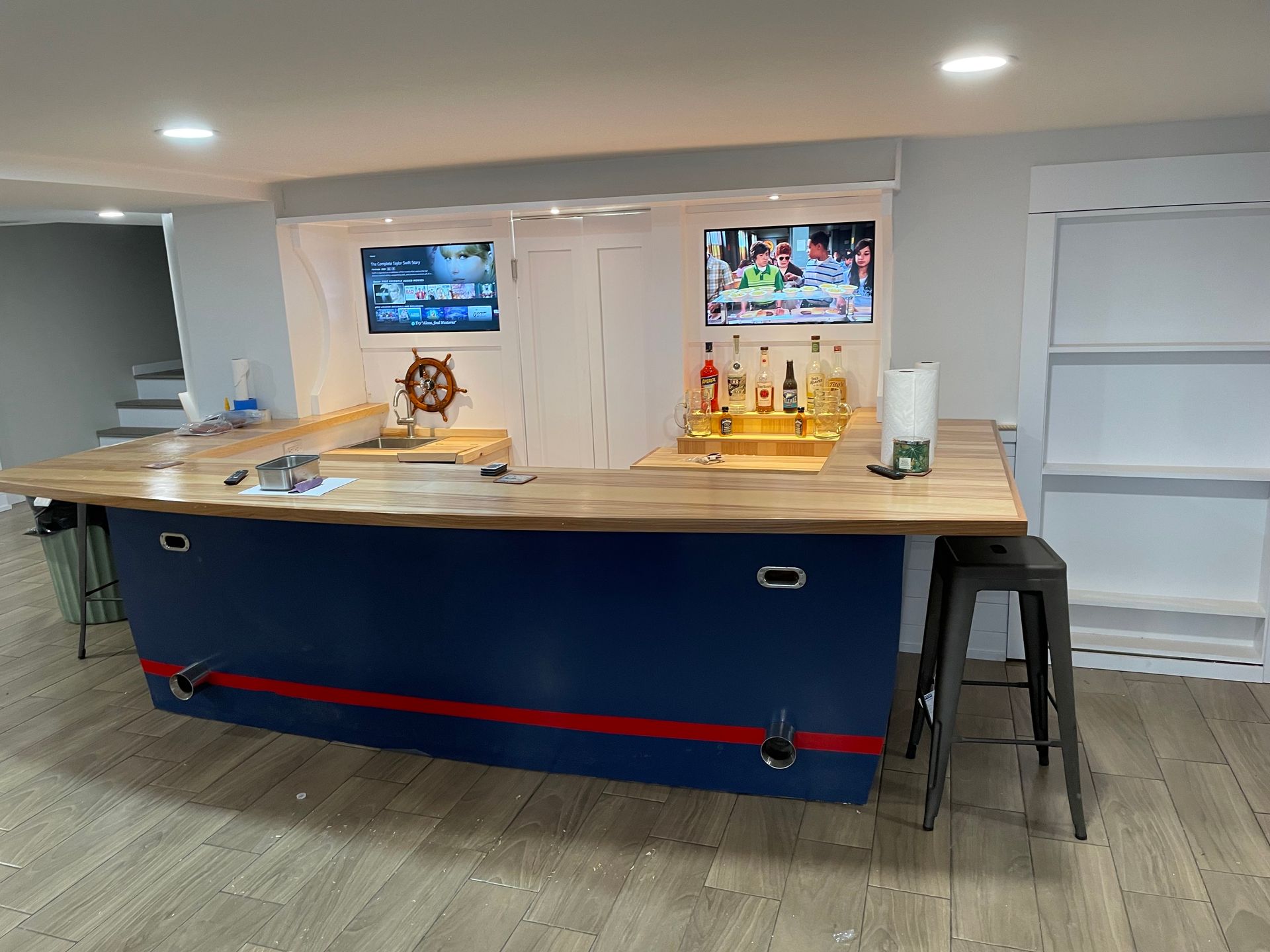 Home bar area with blue counter, wooden top, stools, and two wall-mounted TVs.