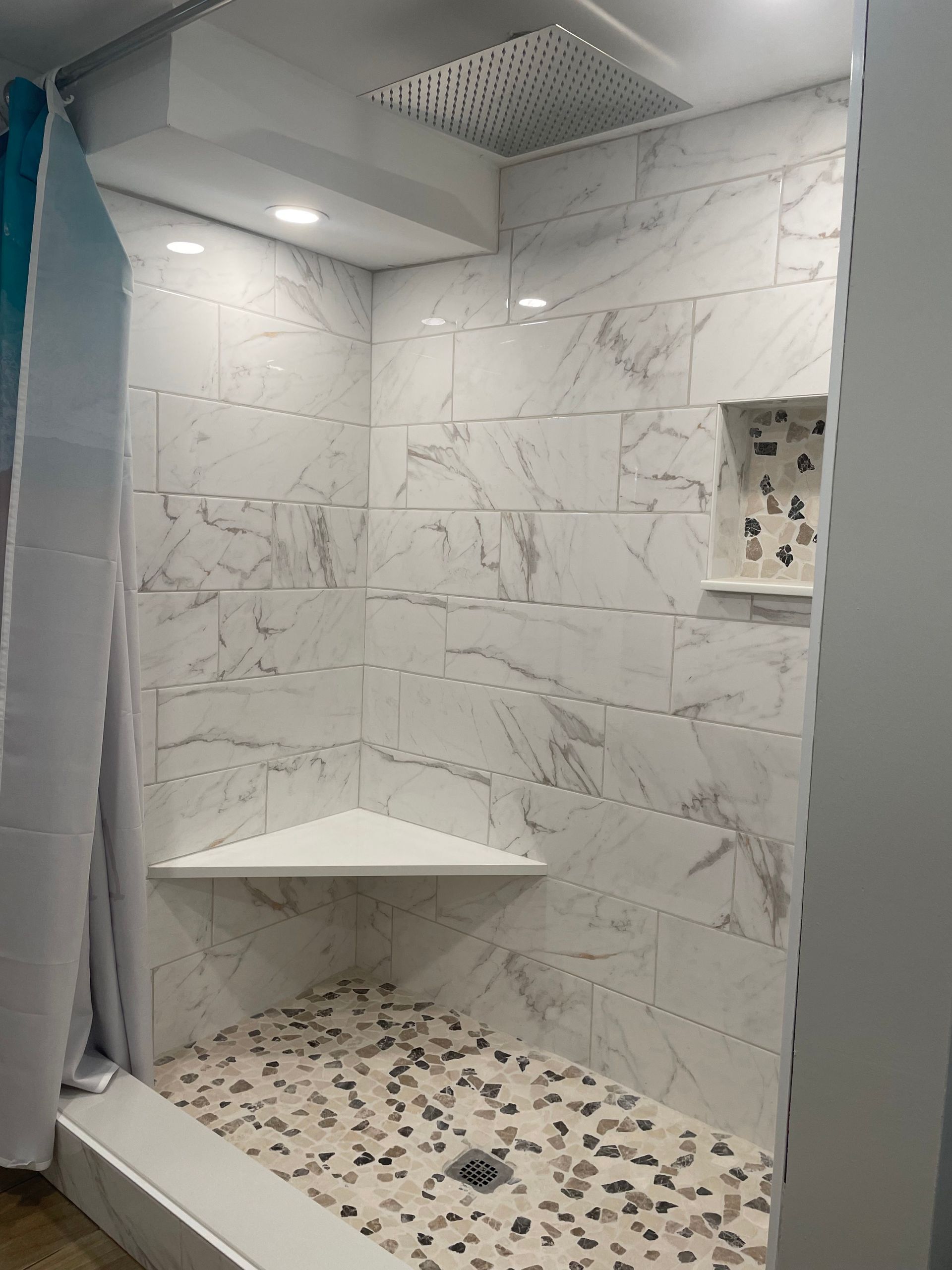 Modern walk-in shower with marble-look tile, corner shelf, and pebble mosaic floor.